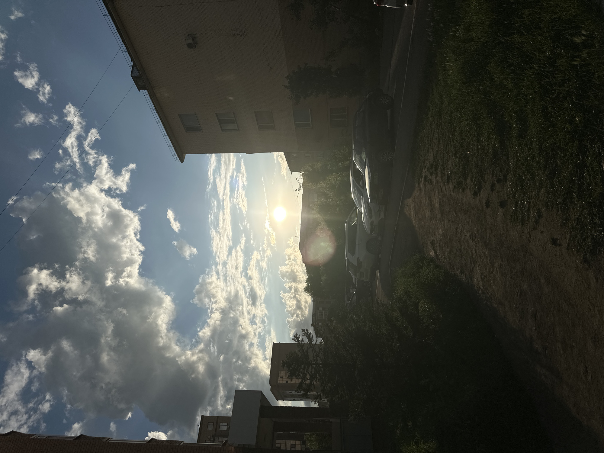 The image depicts a street scene in Kokshetau, Kazakhstan, during what appears to be late afternoon or early evening. The sun is low in the sky, casting a warm glow and creating lens flare. Several apartment buildings of varying heights are visible, constructed with brick and concrete. Some buildings have visible windows, air conditioning units, and balconies. The foreground features a dirt path bordered by grass and a dense green bush. Several cars are parked along the street, including a dark-colored sedan and a white SUV. The sky is a mix of blue with scattered clouds, some of which are illuminated by the sun. The overall impression is a residential area bathed in the golden light of the setting sun.