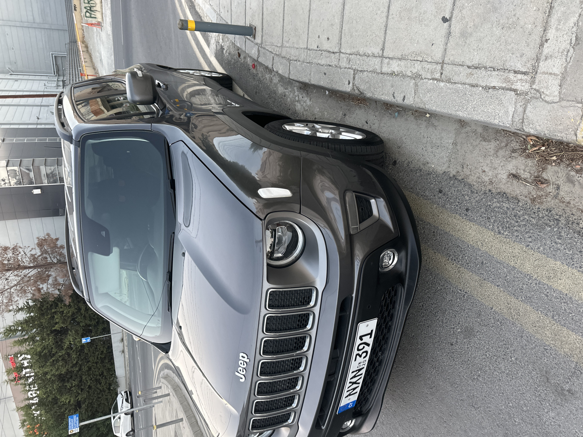 A dark gray Jeep Renegade is parked on the side of a street in an urban environment. The car is facing towards the viewer at a slight angle, with its front end and driver's side visible. The vehicle has a license plate that reads "CY NXN 391". The Jeep is positioned next to a concrete sidewalk with a curb, and a yellow-topped bollard is standing on the sidewalk. The street appears to be paved with asphalt and has yellow lane markings. In the background, there is a building with a modern facade and some trees. Further in the distance, another car is visible on the street. The scene appears to be in daylight, and the lighting suggests it is a clear day.