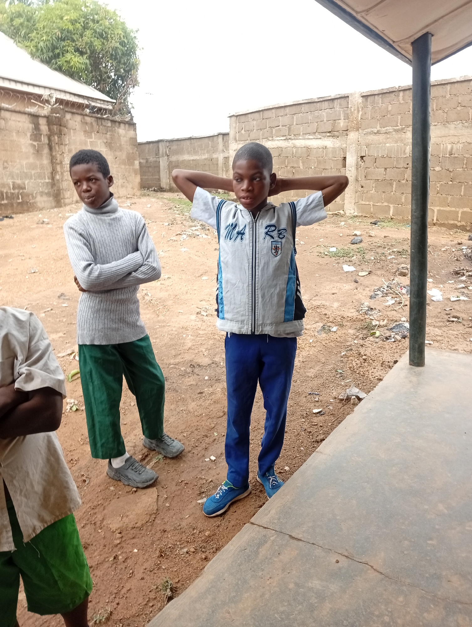 Three young boys are standing outside. The boy on the left is wearing a gray turtleneck sweater and green pants, with gray shoes. The boy in the middle has his hands behind his head and is wearing a gray and white jacket with blue accents, blue pants, and blue shoes. The boy on the left side of the image is wearing a tan short-sleeved shirt with green pants. The setting appears to be outdoors in Narayi, Nigeria, with a building behind them. The ground is dirt with trash strewn about.