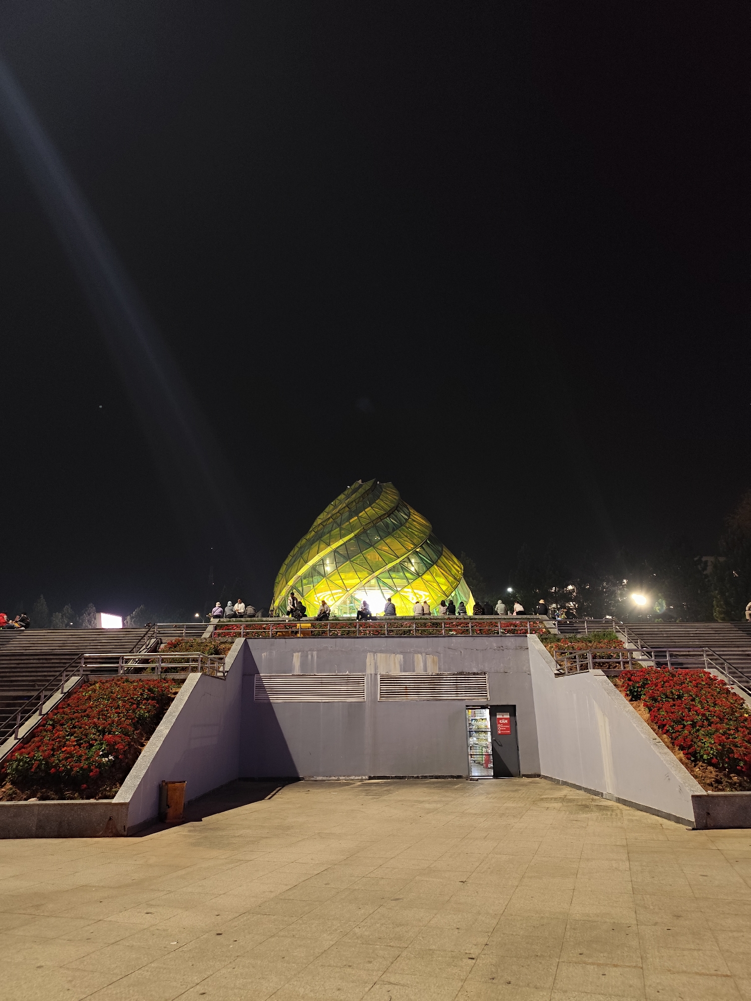 This nighttime image depicts the Da Lat Flower Park Exhibition Hall, a distinctive spiral-shaped glass structure illuminated with yellow-green light, situated in Da Lat, Vietnam.
The scene is viewed from a ground-level plaza of light-colored paving stones. In the immediate midground, a concrete multi-tiered structure rises, featuring wide staircases on both the left and right flanks. These stairs are bordered by metal railings. Between the staircases and along the base of the elevated platform, sloped landscaped beds are filled with dense red flowering plants.
A central grey concrete retaining wall forms the base of the elevated platform. On the right side of this wall, a dark doorway is visible, revealing an illuminated interior, possibly a shop, with items on shelves and a red sign near the entrance. A yellow-orange trash receptacle stands on the ground level to the left of the doorway.
On the elevated platform and the upper sections of the staircases, approximately a dozen individuals are scattered, either standing or seated, facing towards the illuminated exhibition hall. They appear to be visitors or patrons. The Da Lat Flower Park Exhibition Hall itself dominates the upper midground, glowing brightly against a dark, starless sky. Beyond the structure, the silhouettes of trees and distant artificial lights indicate a surrounding urban environment. A prominent light streak, consistent with a lens flare, extends diagonally from the upper left corner of the frame across the dark sky.