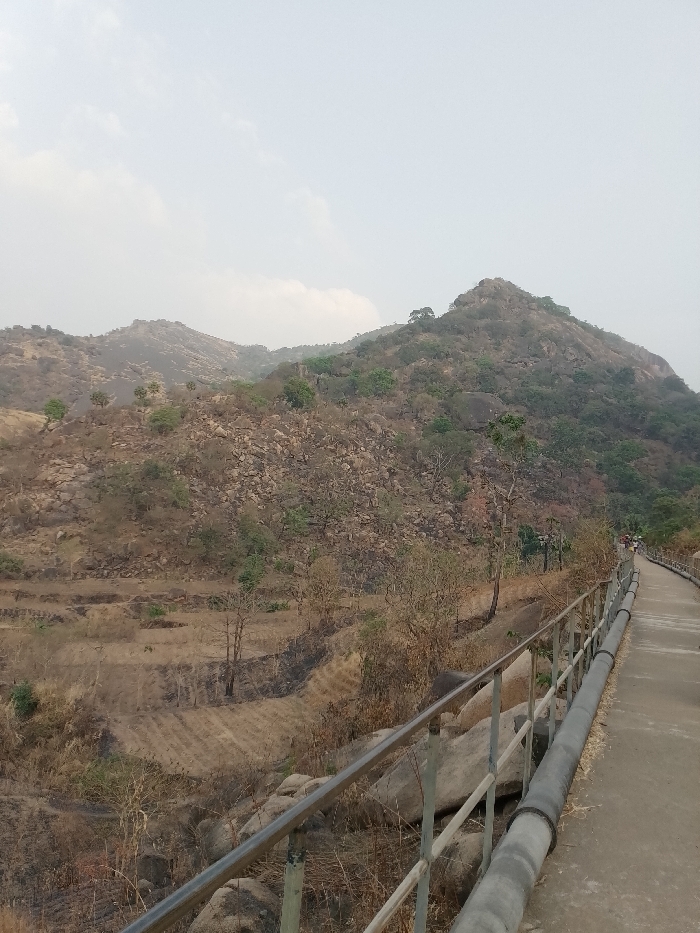 Hills dominate the background, sparsely vegetated with trees and rocks. A concrete pathway stretches into the distance, lined with a metal railing and a large pipe running along its side. Barren, terraced fields and scrubby vegetation fill the area between the path and the hills. A hazy sky creates a muted, overcast light.