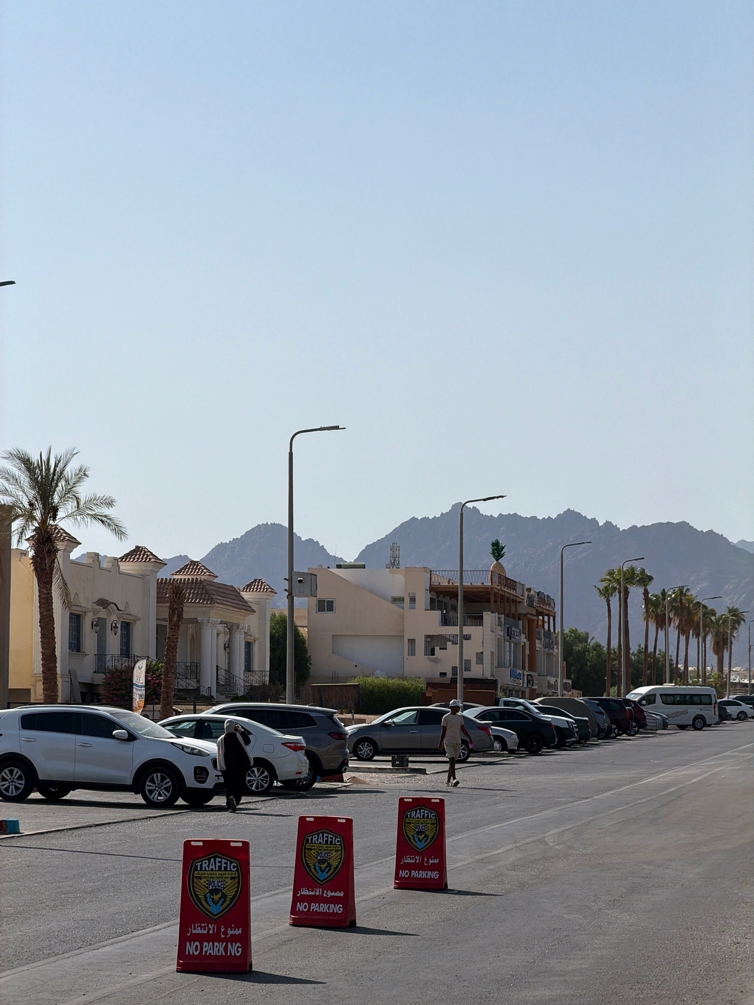 This is a street scene in Sharm El Sheikh, Egypt. The image shows a row of parked cars along a street, with buildings and palm trees on the left side and mountains in the background. Two red "NO PARKING" signs are visible in the foreground. A few people are seen walking on the sidewalk and near the cars. The sky is clear and blue. The architecture of the buildings suggests a resort or residential area. The overall impression is a typical sunny day in a coastal city.