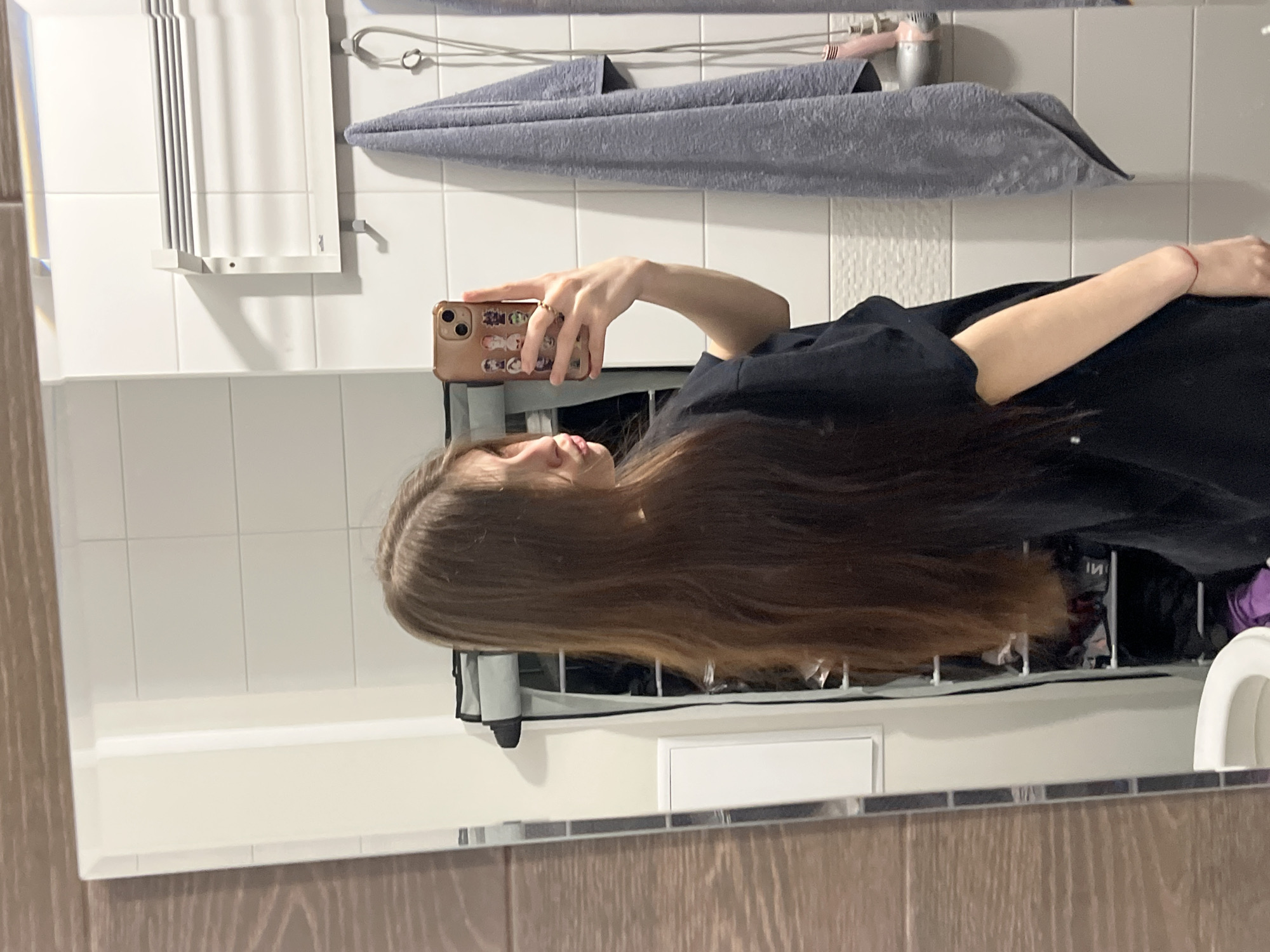 A young woman with long, brown hair is taking a mirror selfie in a bathroom. She is wearing a black t-shirt and holding a gold-colored smartphone with stickers on the back. The bathroom has white tiled walls and a dark wood-look vanity. A towel rack with a gray towel and a hair dryer attached is visible. A white radiator hangs on the wall. A toilet is visible in the lower right corner of the mirror reflection. The location is most likely a private residence. There is no information to confirm the location is in Saint Petersburg, Russia.