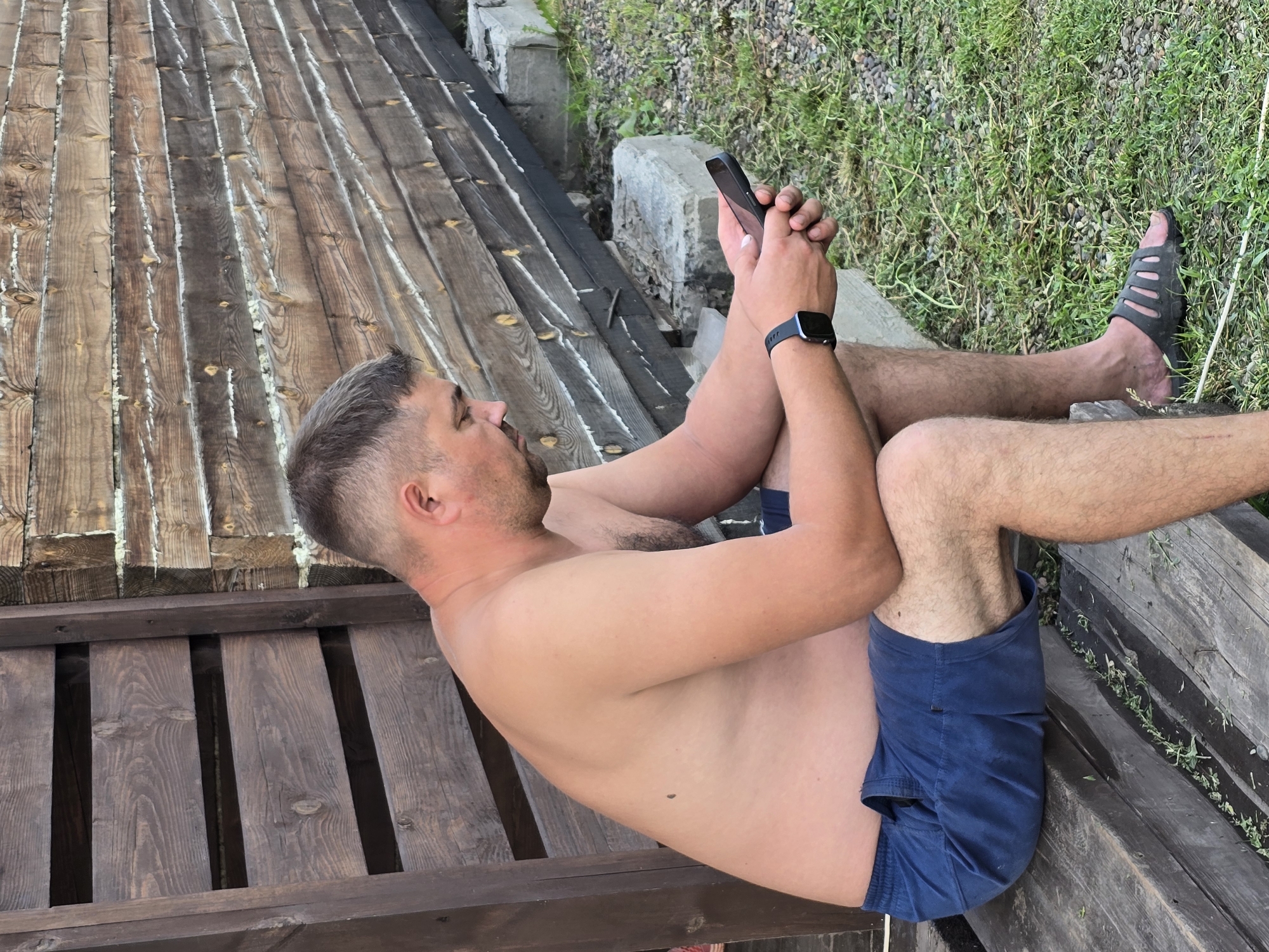 A shirtless man in blue shorts and a black sandal sits on a wooden bench, looking at his phone. He has short, dark hair and a light complexion. Behind him is a rustic wooden structure with horizontal planks and a rough, textured finish. The ground is covered in dry grass and small stones. The scene appears to be outdoors, possibly in a rural or semi-rural setting.