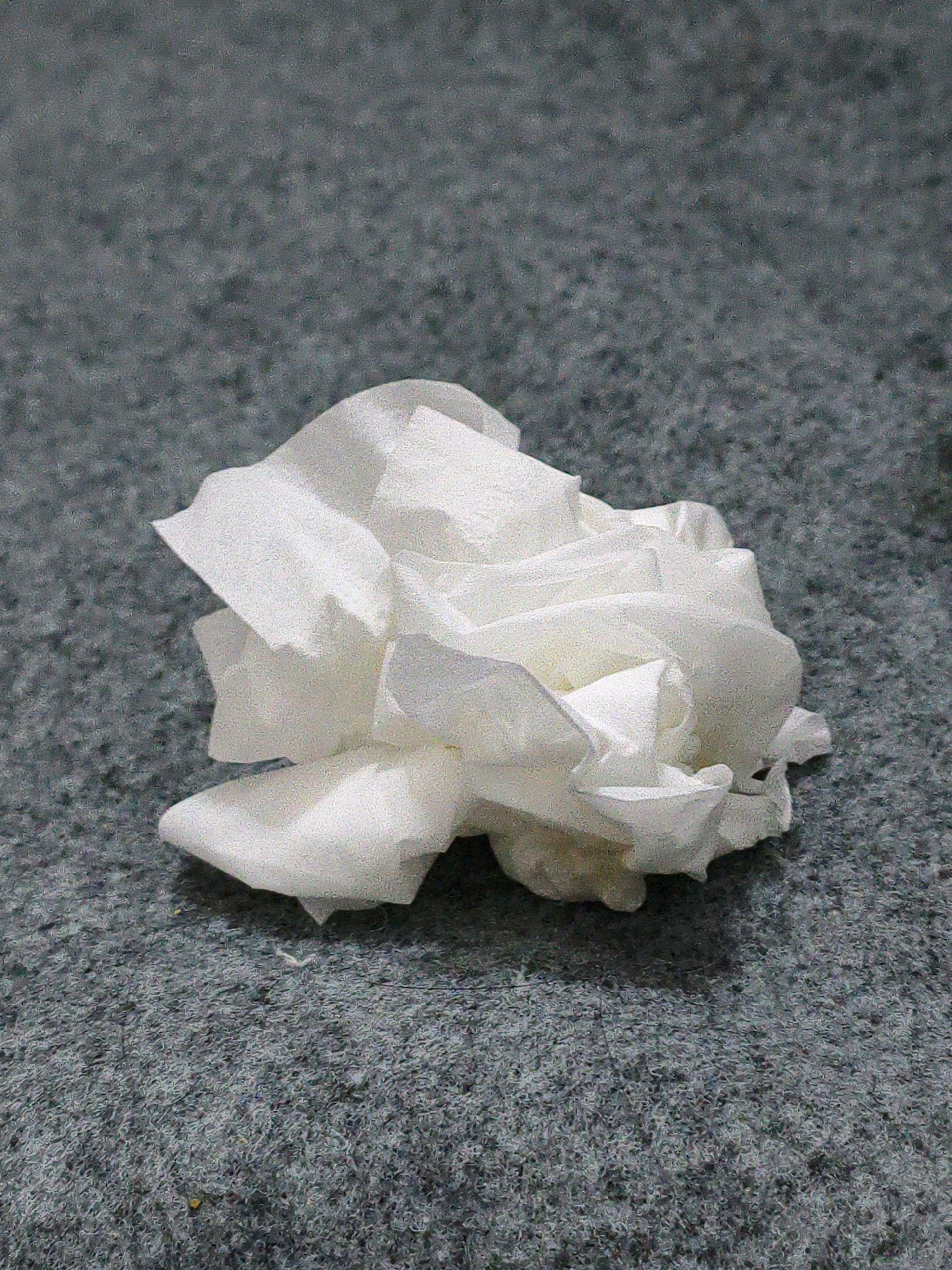 A crumpled white tissue is resting on a gray carpet. The tissue is positioned slightly off-center to the right. The lighting appears to be from above, casting subtle shadows around the tissue. The carpet has a visible texture and appears to be of a neutral gray color. There are no people or other objects in the scene. The image quality has a grainy texture. No information is available regarding the location in the city of Karangtalun, Indonesia.