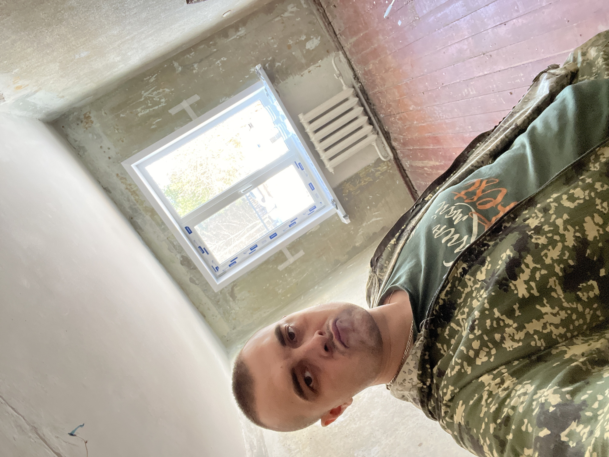 A male individual is depicted in a close-up, high-angle selfie within a room undergoing renovation, located in the city of Слобода, Russia. The man, with short buzzed hair and a direct, slightly pouting expression, wears a green camouflage hooded jacket over a dark green t-shirt with orange text, and a thin gold-colored chain. The room's walls are stripped, revealing light greenish-grey plaster with peeling sections and remnants of old finishes, along with patches of white tape. A modern, white PVC-framed double-pane window is installed on the right, providing natural light and showing a view of bare tree branches outside. Below the window is a white, ribbed cast-iron radiator. The floor consists of worn, reddish-brown wooden planks with scattered debris. The ceiling is white and relatively smooth, with a blue electrical wire visible.