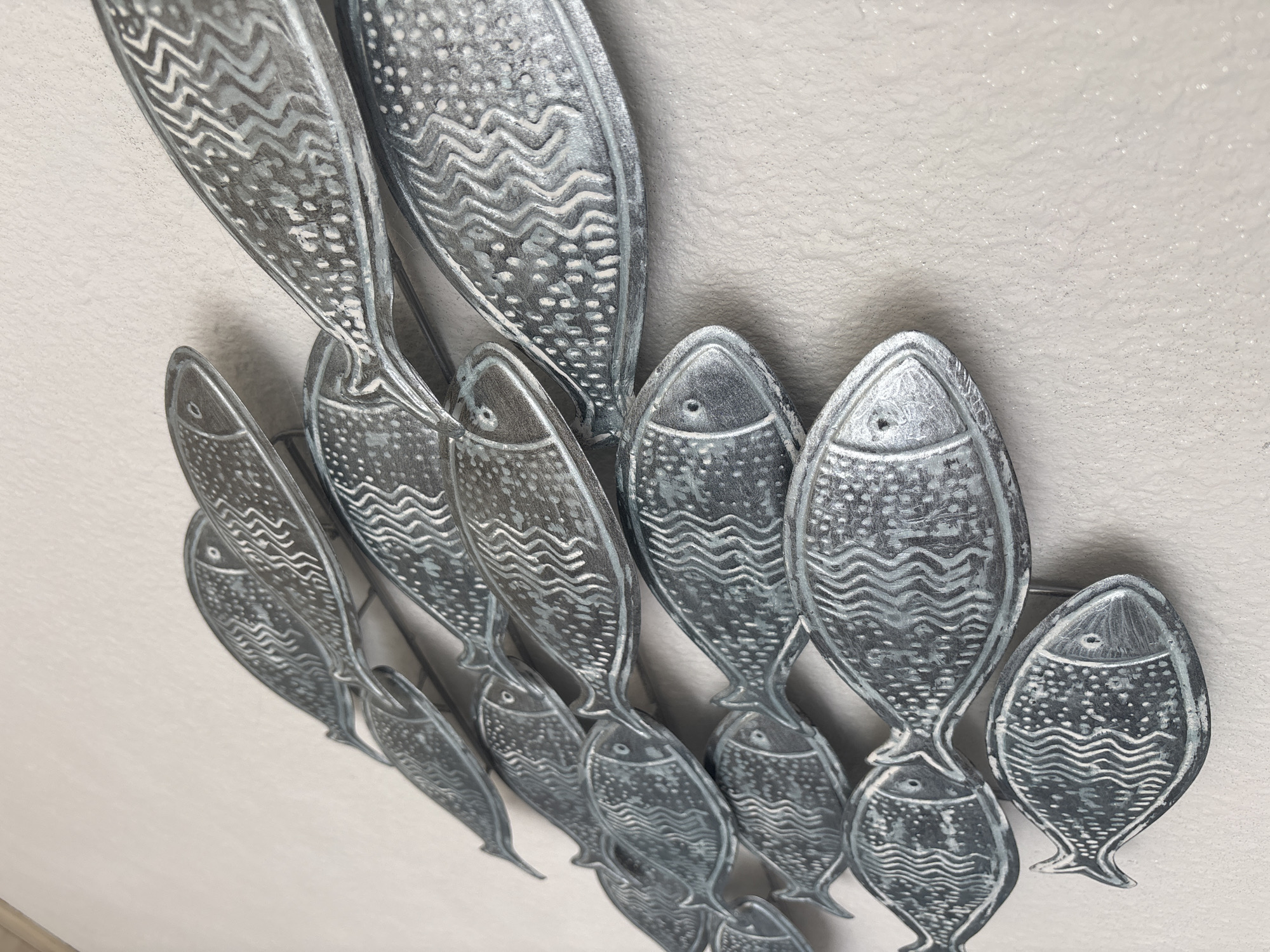 A decorative metal wall art piece depicting a school of stylized fish is mounted on a textured, off-white wall. The fish are arranged in a diagonal line, moving from the upper left to the lower right of the frame. Each fish is metallic gray with lighter gray or white decorative markings, including wavy lines along the body and dots. The lighting creates subtle shadows, highlighting the three-dimensional quality of the artwork. The background wall has a slightly grainy texture with fine sparkles. The specific location in the city of Unknown, Unknown is not discernible from the image.