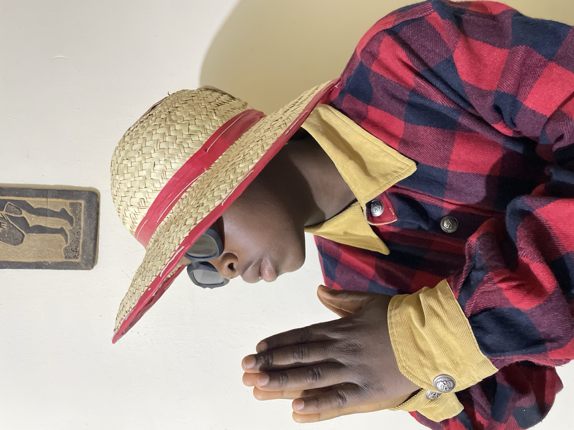 An individual is depicted indoors, wearing a straw hat with a red brim and dark sunglasses. The person is also wearing a red and black plaid shirt with a tan corduroy collar and cuffs, which are secured with silver buttons. The person has dark skin, and their hands are clasped together. A framed, carved object hangs on the wall in the background. There is no verifiable information about the location being in Jos, Nigeria, in the provided image.