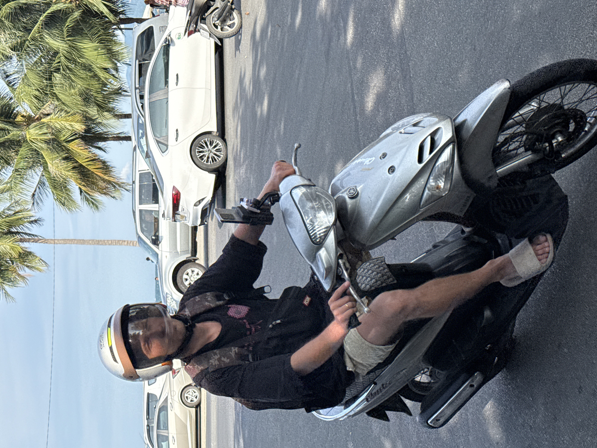 A young man wearing a silver helmet and black t-shirt rides a silver scooter on an asphalt road. He has a camouflage backpack on and is wearing tan shorts and light-colored sandals. Palm trees line the background, suggesting a tropical location. Several cars are parked or driving in the background. The sky is bright blue with some white clouds. The image appears to be taken during the daytime.