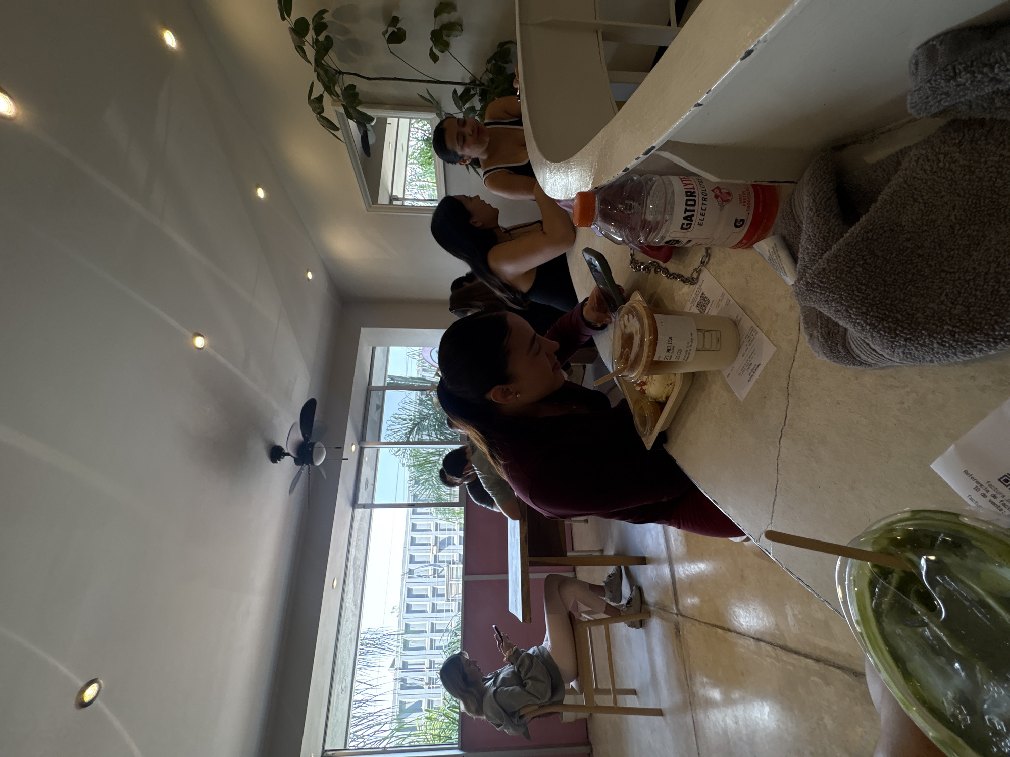 The image captures a well-lit interior, possibly a cafe or restaurant, where several individuals are present. A woman with dark hair, wearing a burgundy top, sits at a counter and appears to be interacting with her phone, next to a cup of coffee with her name. The counter also features a bottle of Gatorlyte, a receipt, a gray towel, and a plastic cup filled with a green liquid and a straw. Further down the counter, two other women are seated, facing the opposite direction. Additional people can be seen in the background, seated at tables. A woman near a window, sitting on a chair and also interacting with her phone. Outside the windows, buildings and greenery are visible, suggesting an urban setting. A ceiling fan and recessed lighting are visible on the ceiling. Based on the presence of a receipt containing Spanish words, it is possible that the location is in Puebla, Mexico.