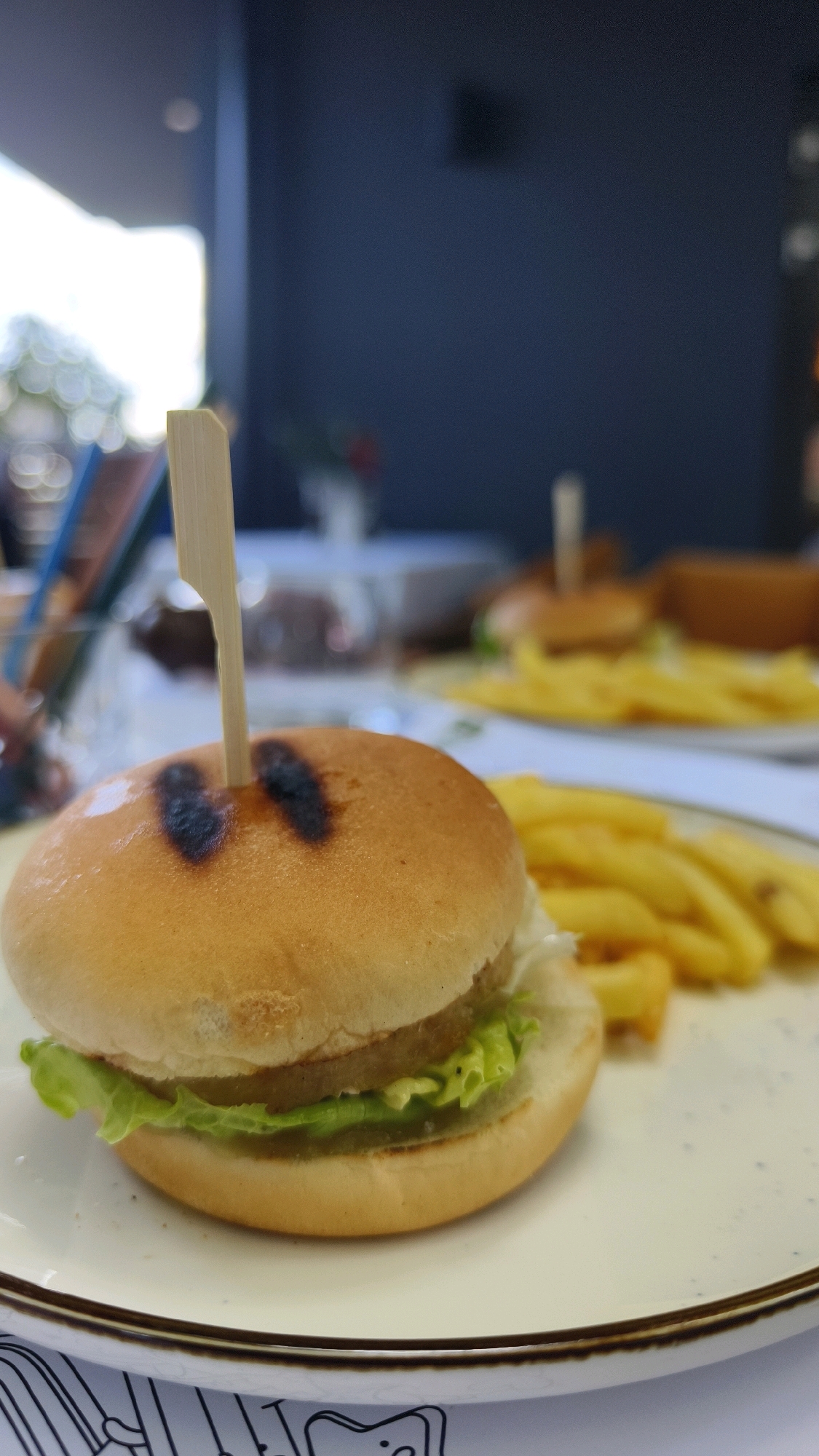 The image shows a close-up of a mini burger on a white plate, with French fries in the background. The burger has a wooden skewer inserted through its top bun, which is slightly charred. Fresh green lettuce is visible peeking out from the bottom of the burger. The scene appears to be set at an outdoor cafe or restaurant, possibly in Vienna, Austria, given the blurry background which suggests an urban environment with foliage. The lighting indicates it is daytime. There are no people visible in the frame, and no actions are taking place. The focus is solely on the food presented.