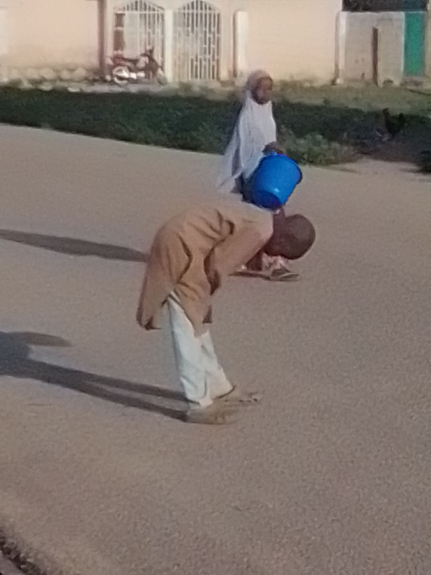 In Gombe, Nigeria, a person wearing a brown tunic and white trousers is bending over, holding a blue bucket. Another person, dressed in a white headscarf and garment, stands in the background holding a similar blue bucket. A motorcycle is parked near a building with white barred doors in the background, and a chicken is visible on the grass. The scene is set on a paved road.