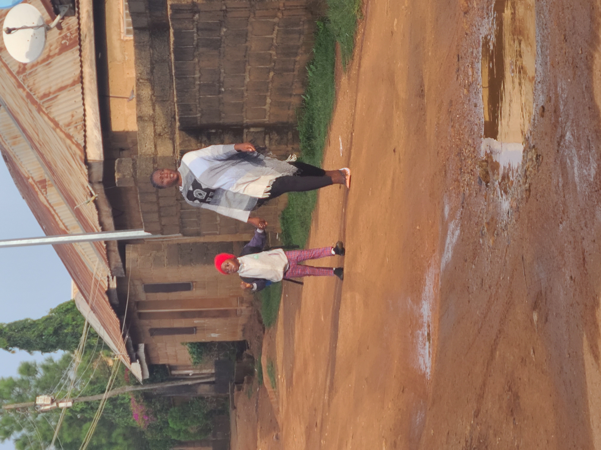 The image depicts two people walking on a dirt road in Jos, Nigeria. An adult, dressed in a white patterned shawl and dark pants, walks beside a child wearing a red head covering, a light-colored top, and checkered leggings. The road appears rutted with a large puddle reflecting the sky and surrounding structures. The environment includes buildings constructed of concrete blocks and corrugated metal roofing. Vegetation is visible in the background and along the roadside. Electrical wires can be seen strung between poles. The overall atmosphere suggests a rural or semi-rural area within the city.
