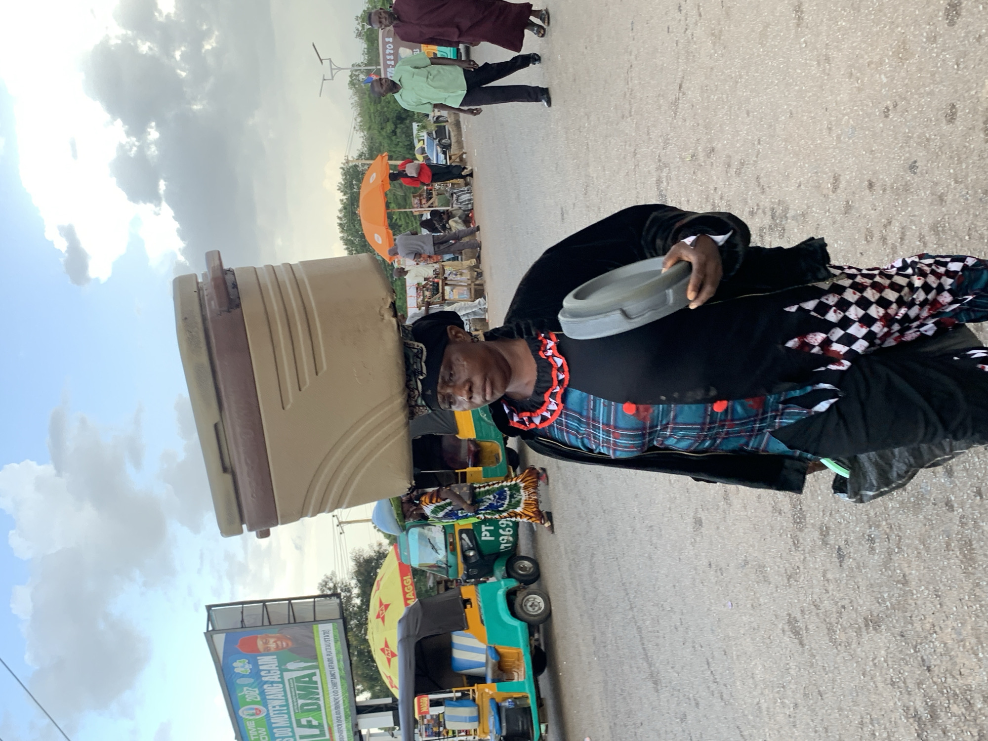In Jos, Nigeria, a woman walks down a street carrying a large cooler on her head and a lid in her right hand. The woman is wearing a black jacket over a patterned top and black pants with a checkered pattern at the hem. She also wears a black headscarf and a beaded necklace. In the background, several auto-rickshaws, commonly known as "keke napep," are parked or in motion. A billboard advertising a political candidate, with the text "LET THE TIME COME. 2023. LET US DO MUTFWANG AGAIN," is visible. The sky is partly cloudy, and the scene suggests a typical street market or transportation hub.