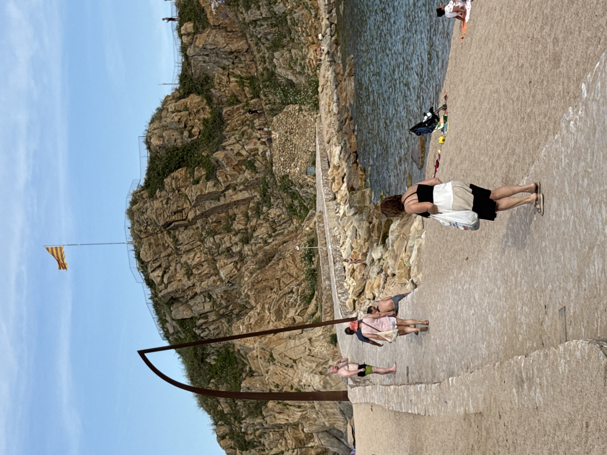 The image captures a scene along a paved walkway that hugs a rocky coastline. In the background, a rocky hill is visible, topped with a Catalan flag flying from a flagpole. A metal sculpture resembling an arch rises from the left side of the walkway.
Several people are present on the walkway and the adjacent sandy beach. On the walkway, a man in swim trunks stands facing away from the camera, while a group of three individuals, two women and a man, walk away from the viewer. Further along the walkway, a woman wearing a black and white outfit walks towards the camera, carrying bags. On the beach, a few individuals are seen relaxing, including a woman sitting on a towel and another person lying down. A baby stroller and other beach accessories are scattered on the sand. The sea is to the right, with gentle waves lapping against the rocky shore. The environment appears to be a coastal area in Blanes, Spain, likely a popular spot for recreation.