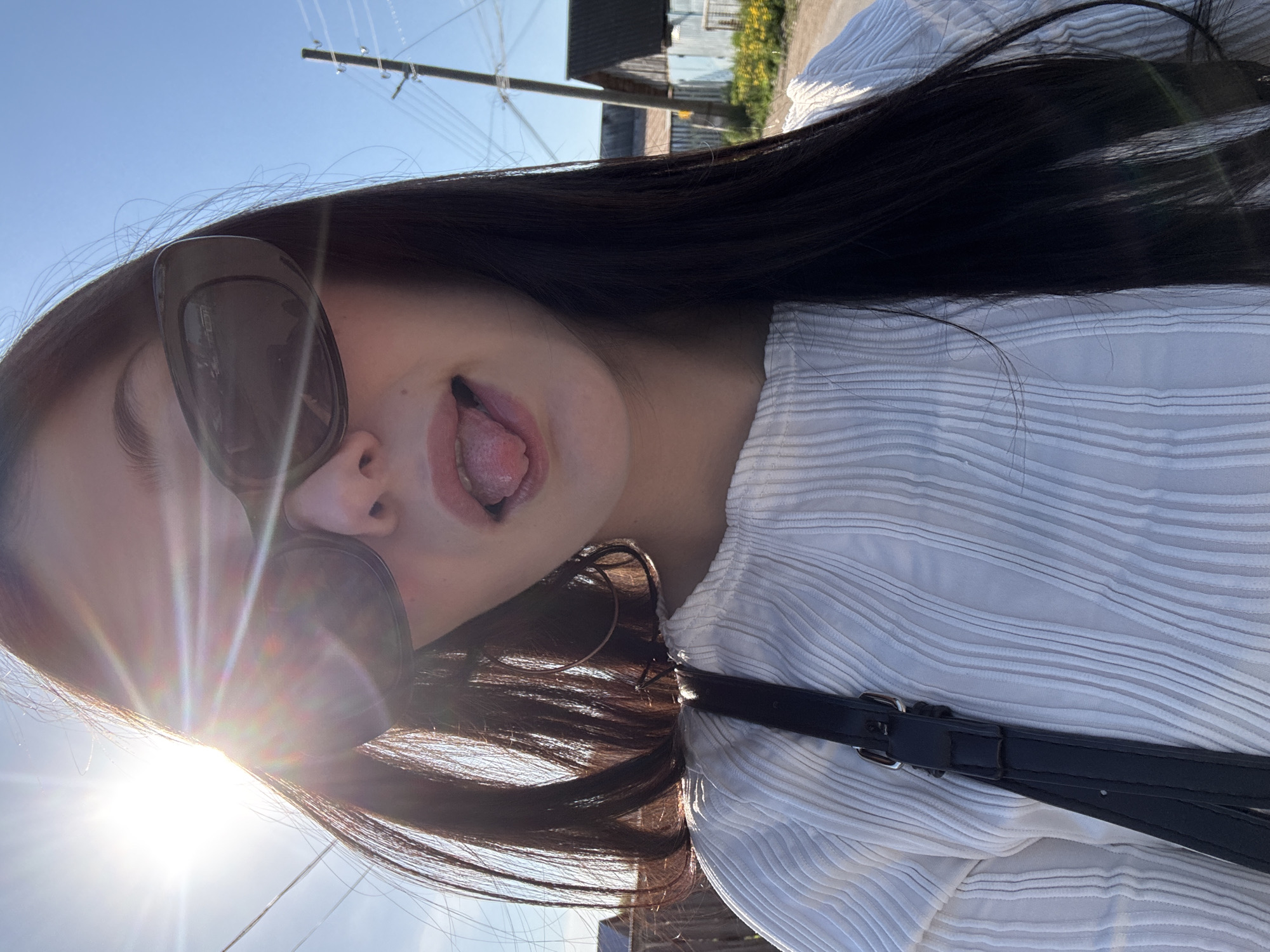 The image shows a woman with long, dark hair wearing sunglasses and a white shirt with horizontal stripes. She is sticking her tongue out. A black strap, possibly from a purse or bag, is visible across her chest. The background shows a clear blue sky, power lines, and a glimpse of buildings and greenery. The bright sunlight suggests it is daytime. Given the request for location in the city of Фоки, Russia, the environmental elements suggest a residential area with infrastructure. There are no explicit indicators confirming the exact location within Фоки, Russia.