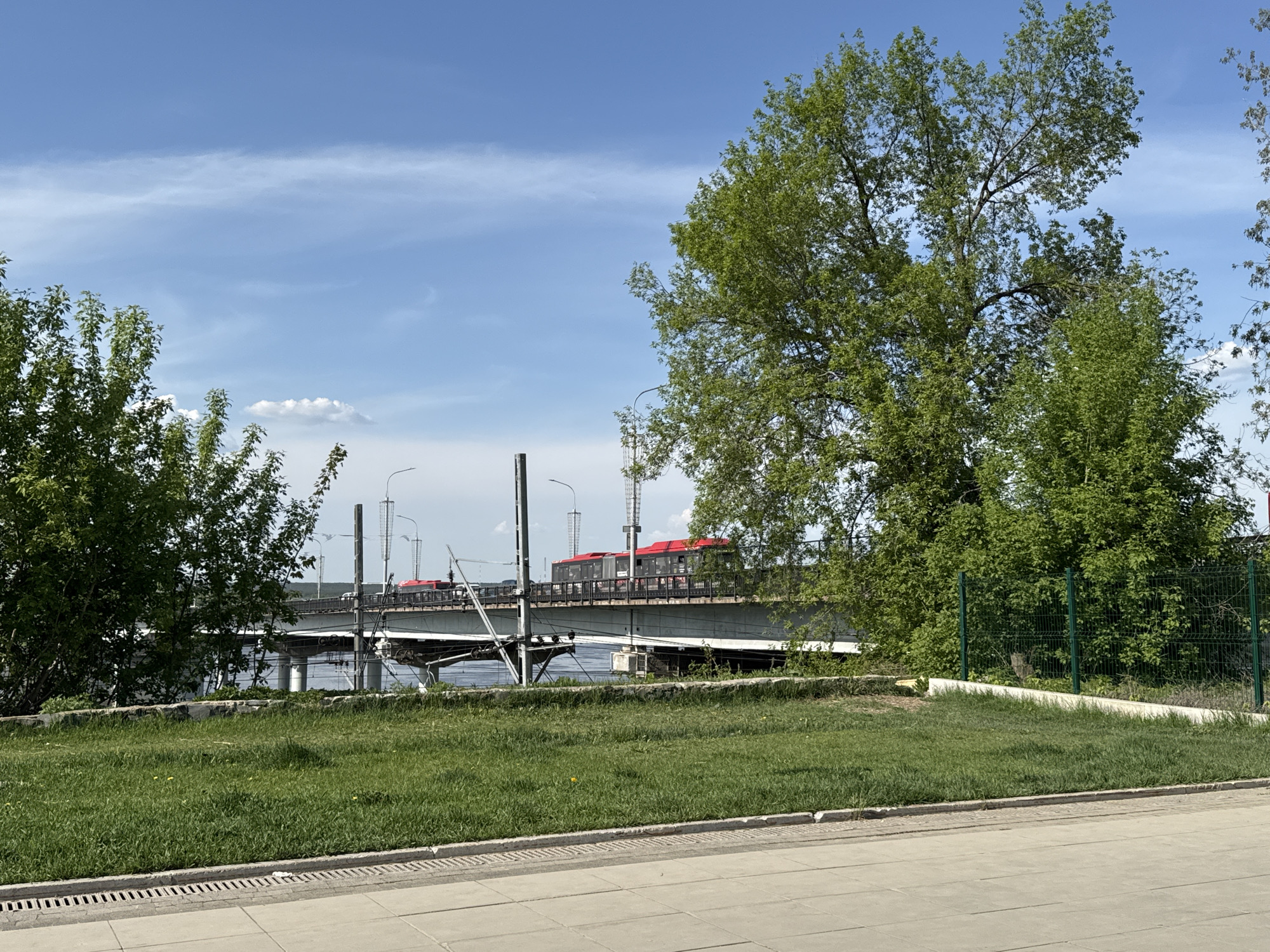The image shows a view of the Kama River Bridge in Perm, Russia. A tram is seen crossing the bridge, which also carries vehicle traffic and streetlights. The foreground consists of a paved area and a grassy bank, partially obscured by trees and shrubs. A green chain-link fence is visible on the right side of the image. The sky is blue with scattered clouds.