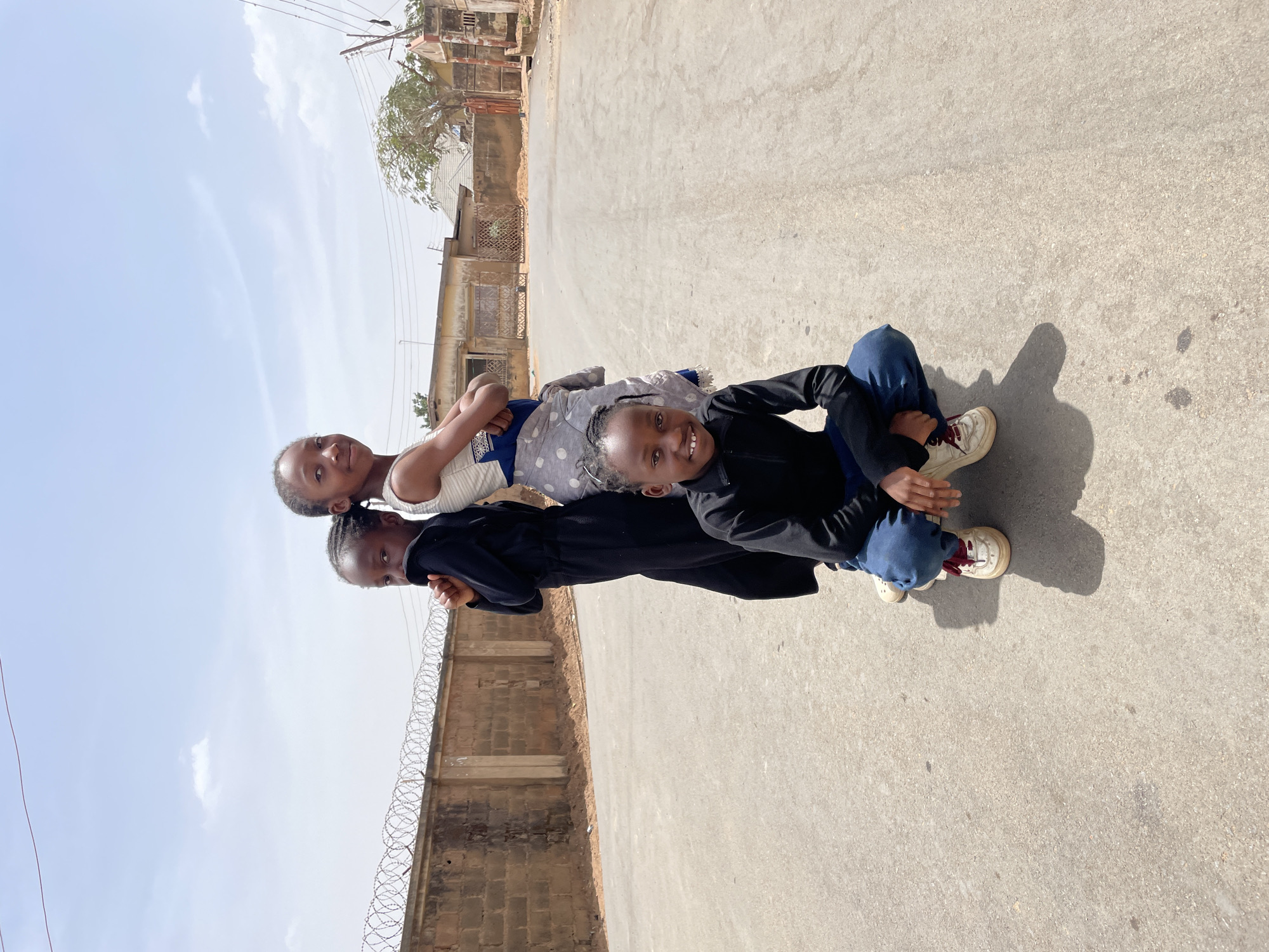A daylight, eye-level photograph shows three children posed on a paved road in Jos, Nigeria. In the foreground, a child, identifiable as male, wearing a black long-sleeved top and blue jeans, squats centrally with a smile directed towards the viewer. He wears light-colored sneakers with red laces. Immediately behind and slightly to his right stands a second child, identifiable as female, wearing a light-colored, patterned sleeveless top and a blue and white patterned wrap garment around her lower body; she faces the viewer with a slight smile, her arms crossed behind her back. To the left and slightly behind the second child, a third child, identifiable as female, stands, turning her upper body to look over her left shoulder towards the viewer; she wears a long black garment and has her left hand raised towards her mouth. All three children have dark skin and braided hairstyles.
The road surface is asphalt or light-colored concrete, showing minor blemishes and scattered dust. To the left of the subjects, running parallel to the road, is a weathered, light brown concrete block wall topped with multiple strands of coiled razor wire. Beyond this wall, a dirt path is visible. To the right, in the background, several single-story, unpainted or light-colored residential buildings with varying rooflines and gates line the road. Utility poles with overhead wires are visible, as well as a large tree with green foliage. The sky above is light blue with scattered white clouds, indicating bright sunlight which casts distinct shadows on the road.