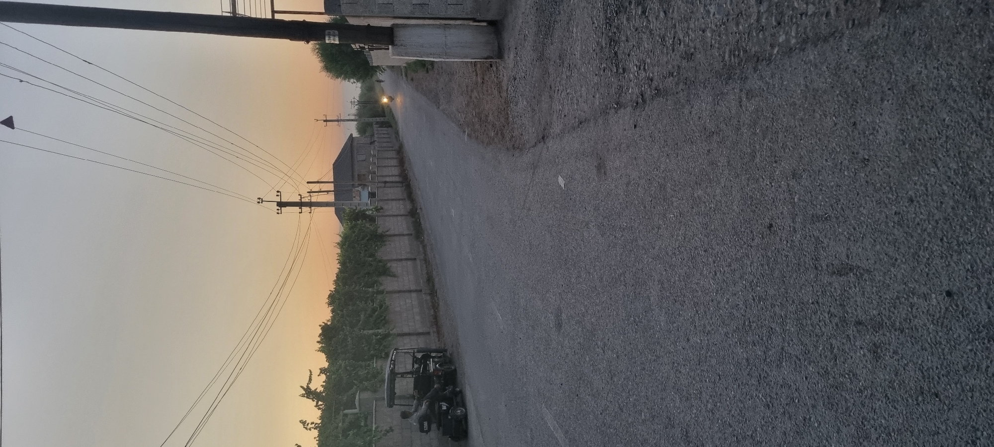 This is a vertical, medium-shot, eye-level, exterior photograph taken in sunlight at dusk. The scene is a rural road in Khovaskor, Tajikistan. The road stretches into the distance, bordered by a stone wall and dense green foliage, likely vineyards. Utility poles with numerous wires are prominent on the left side of the image, silhouetted against a pale orange sky that transitions to a lighter hue towards the top.
In the foreground, a dark-colored, three-wheeled vehicle is parked on the right side of the road. Two individuals are seated within the vehicle, though their features are not clearly discernible. The vehicle appears to be a type of auto rickshaw or tuk-tuk. Further down the road, a motorcycle or scooter is visible, its headlight illuminating the path ahead, suggesting movement.
The overall atmosphere is peaceful and serene, typical of a rural setting at the end of the day. The warm tones of the sunset create a calming visual effect. There is no visible text in the image.