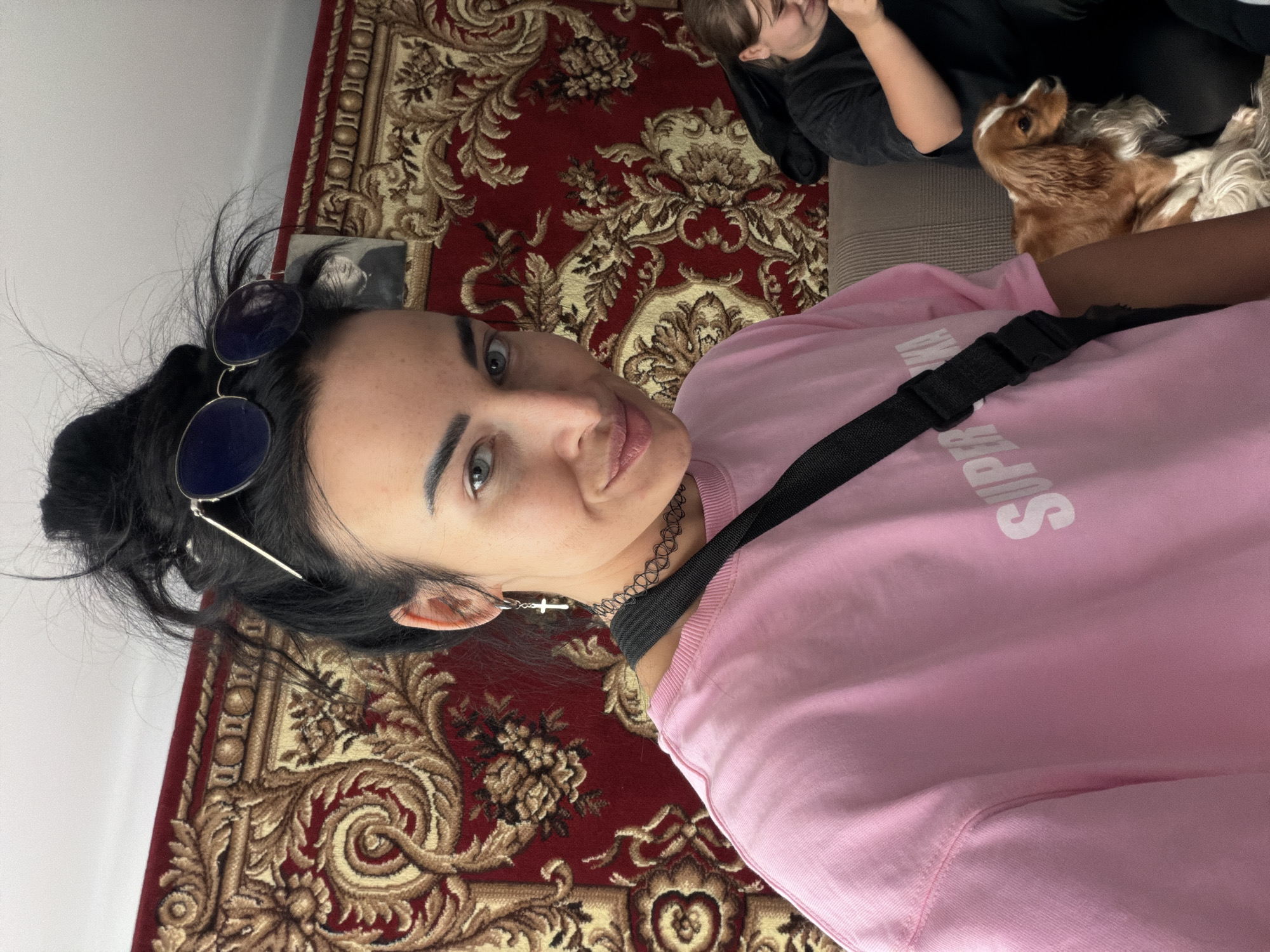A woman is lying on a patterned rug in what appears to be a home interior. She has dark hair pulled back, sunglasses on her head, and wears a pink sweatshirt and a choker necklace. She is looking directly at the camera. To the right of her, a child and a dog are sitting on a couch. The location may be in or near the city of Бронница, Russia.