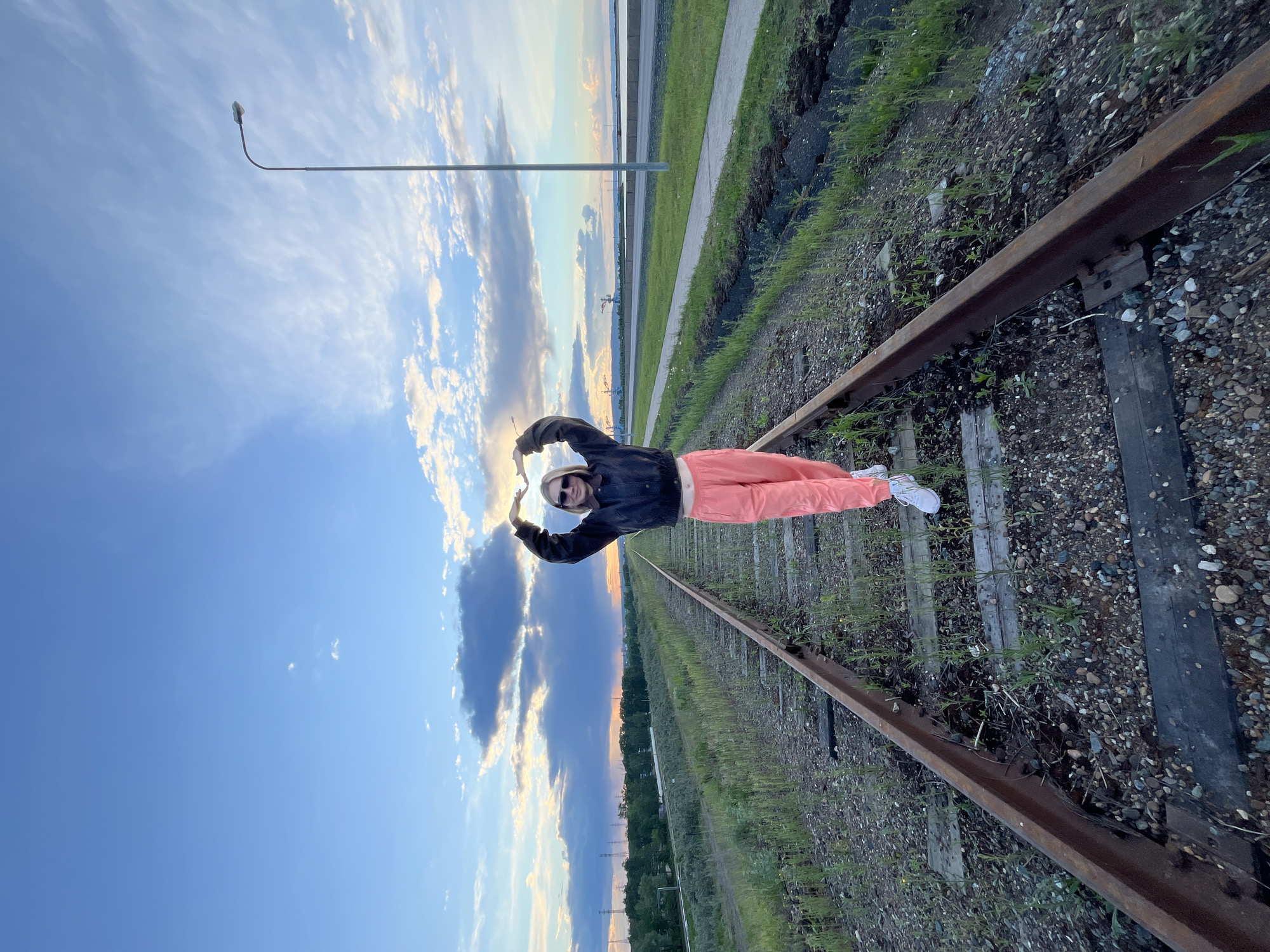 A young person is lying on railroad tracks outdoors. The person is wearing a dark jacket, light-colored pants, and white shoes. The person's arms are raised overhead, forming a partial heart shape around the setting sun. The railroad tracks are bordered by grass and gravel. A lamppost is visible in the background. The location is the city of Chaikovsky, Russia.