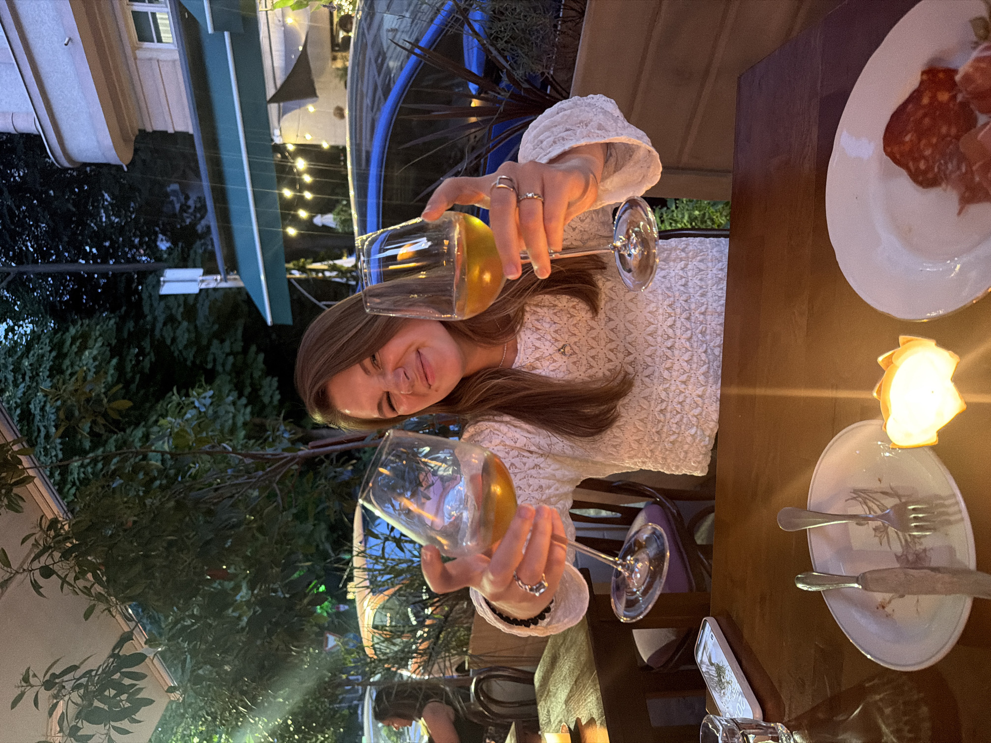 A woman is seated at a table outside a restaurant, holding up two wine glasses filled with a golden-colored liquid. She is making a face, possibly laughing or winking, towards the camera. She is wearing a white, textured sweater and several rings on her fingers. The table is set with plates, cutlery, a phone, and a candle. In the background, there are trees, a building with awnings and string lights, and a car. The overall impression is of a casual evening gathering. The location appears to be a street-side restaurant in Belgrade, Serbia, with the architecture and ambiance suggesting an urban European setting.