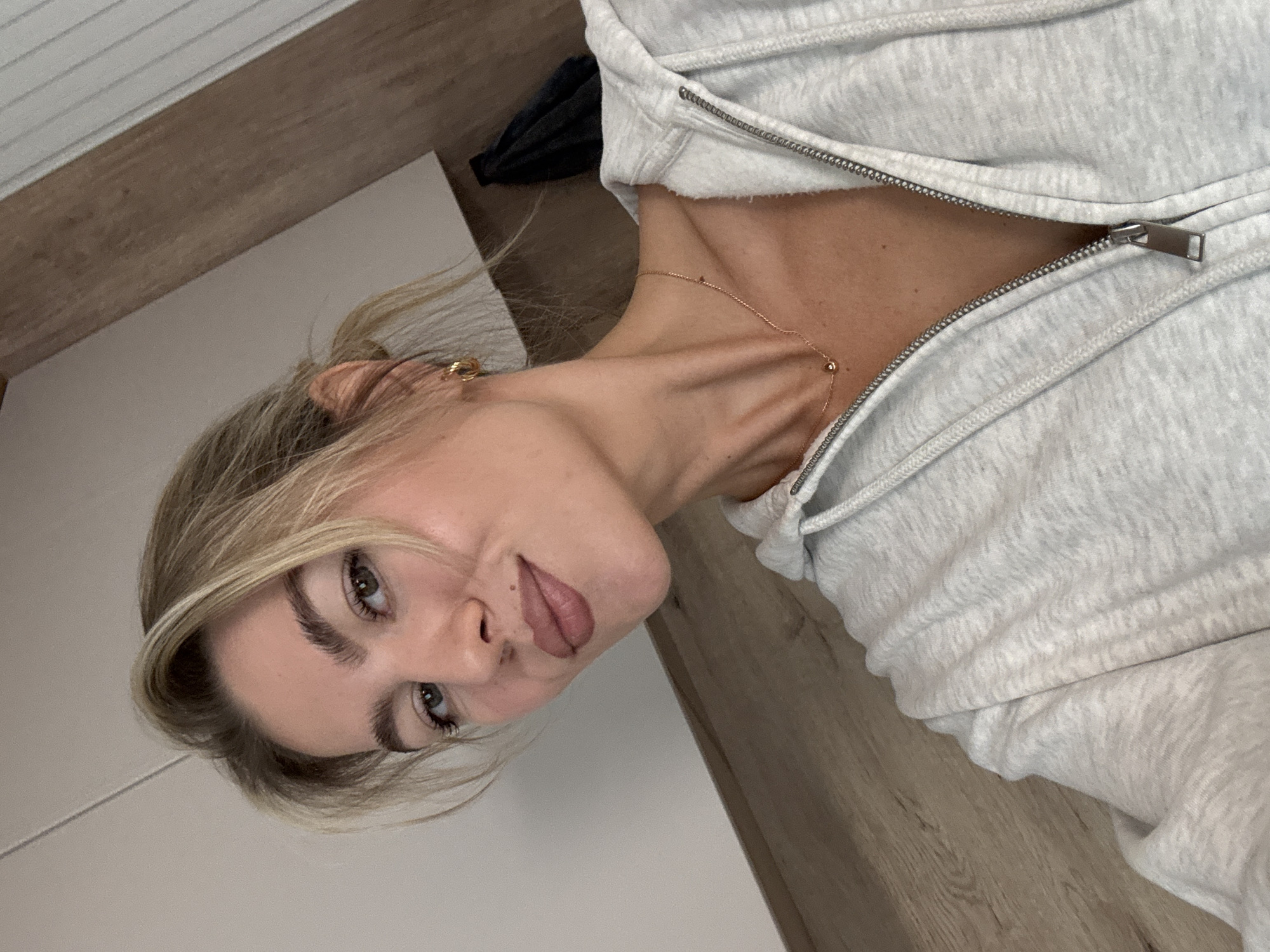 The media file is a close-up, portrait-style photo of a young woman with blonde hair and light-colored eyes. She is wearing a light gray zip-up hoodie, which is partially unzipped, revealing her collarbone and a delicate gold necklace. Her hair is pulled back loosely, with a few strands falling around her face. She has a neutral expression, with her lips slightly parted and her head tilted to the side, looking towards the camera. Her makeup appears natural, with emphasis on her eyebrows and eyelashes.
The background consists of a wooden floor and a white surface, possibly a piece of furniture or a wall. The lighting is soft, suggesting an indoor setting, possibly during the day. There are no other people or objects clearly visible in the frame. The overall impression is casual and intimate, a selfie taken in a personal space. There is no visible text in the image.