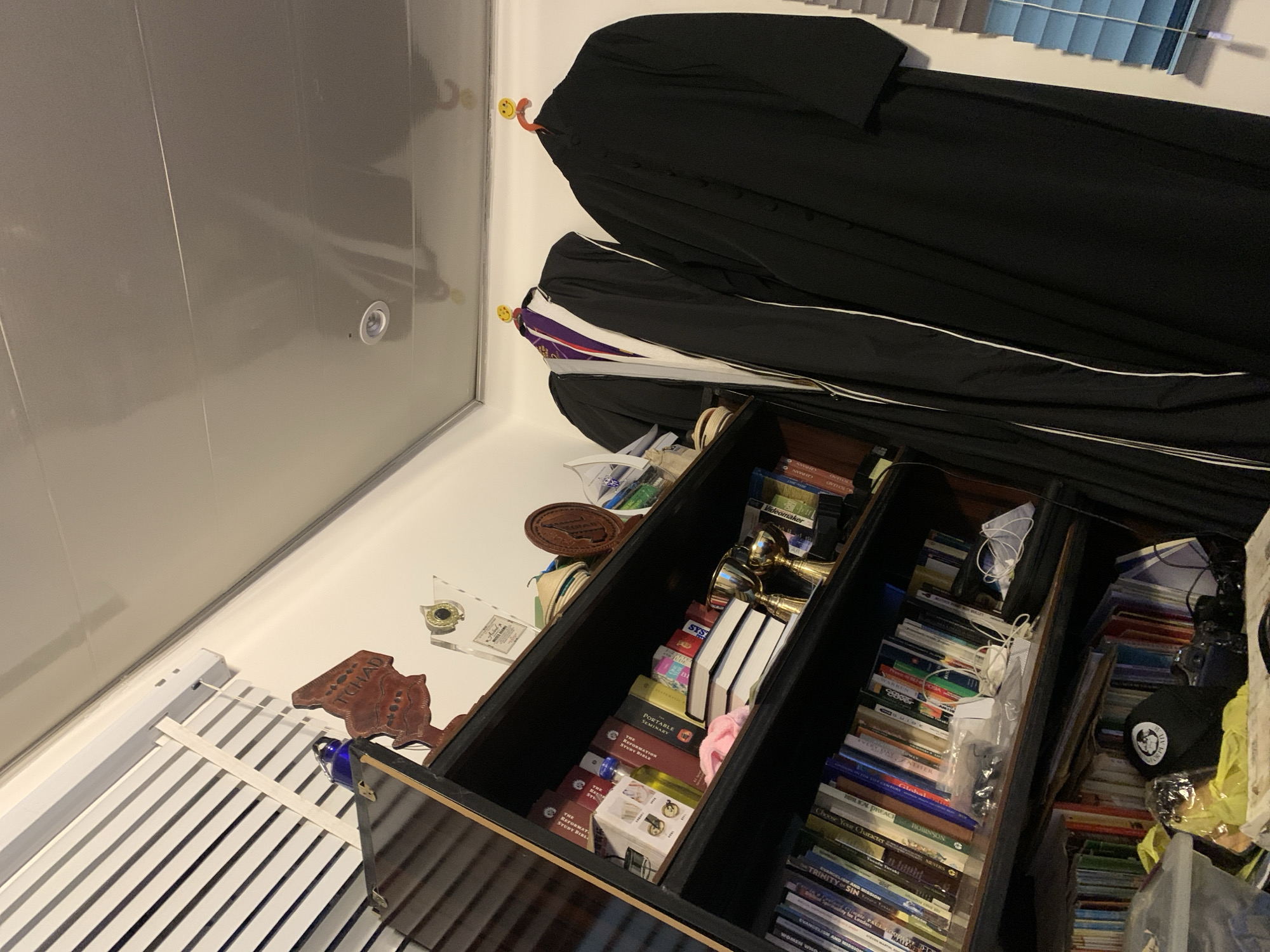 The image shows a cluttered shelf filled with books, trophies, and academic regalia. On the top shelf, a wooden cutout with the word "TCHAD" is visible, along with a blue glass bottle. The middle shelf contains several books, a pink cloth, and two golden trophies. To the right, two academic gowns are hanging on the wall, one with a purple and white sash. The lower shelves are packed with more books and various items, including a black cap with a white logo, and some electronic devices. The overall environment appears to be an indoor space, possibly a study or storage area, with a ceiling that has a reflective surface and a recessed light fixture. There are blinds on a window to the right of the scene. The image is taken from a low angle, looking upwards towards the ceiling.