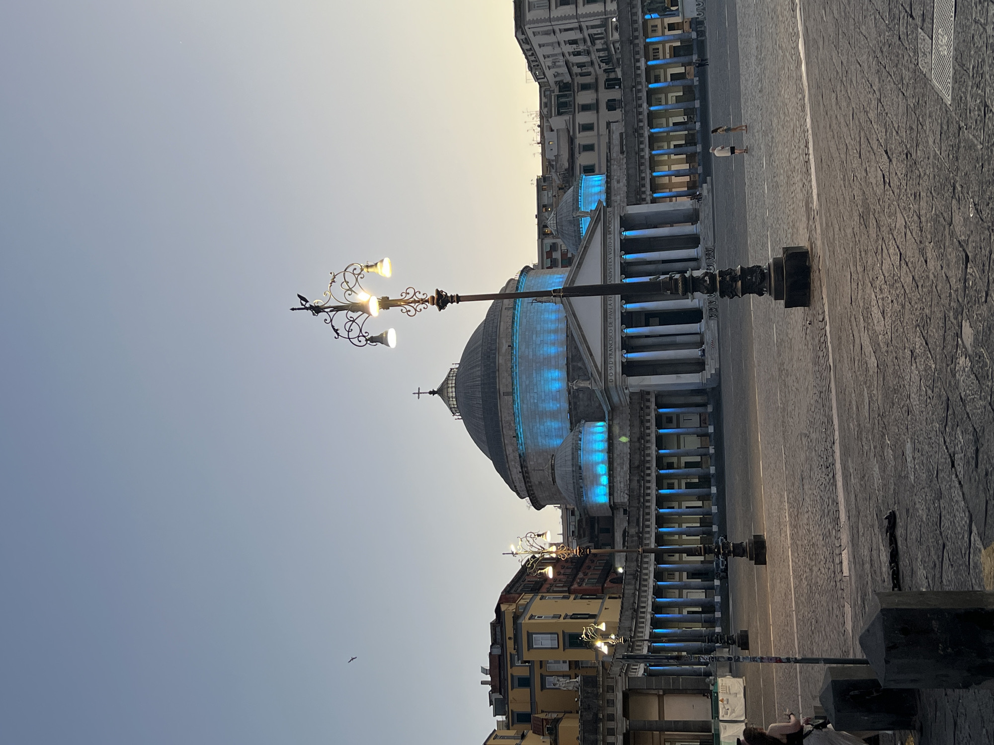 The image depicts Piazza del Plebiscito in Naples, Italy, at dusk. The iconic Royal Palace of Naples, a grand neoclassical building, dominates the background, illuminated by bright blue lights. The palace features a large dome topped with a cross, and a semi-circular colonnade with tall white columns. The cobblestone piazza is vast and empty, with several ornate lampposts casting warm light. A few people are visible in the distance, walking across the square. In the sky, a solitary bird is in flight, and a faint glow indicates the setting sun. The architecture and lighting suggest an evening scene in a well-known public square.