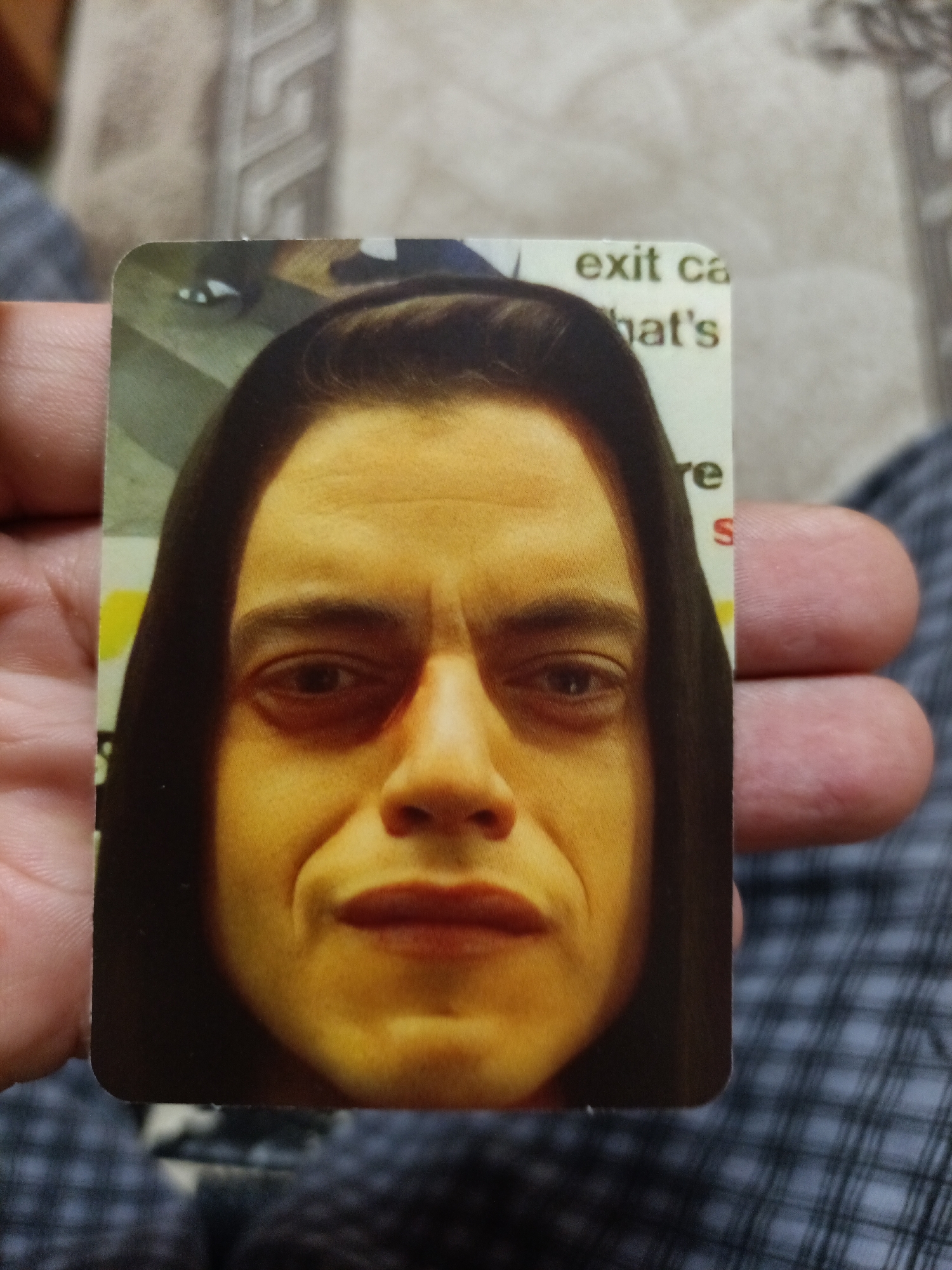 A close-up photograph shows a hand holding a rectangular card featuring a selfie of a man. The man has a dark hooded garment on and his face is lit from the side, creating shadows. His eyes are open, and he has a slight smile. Text is visible on the card, including "exit ca" and "hat's". The background of the photograph is blurred and appears to be a fabric surface. The image was taken in Nyagan, Russia.