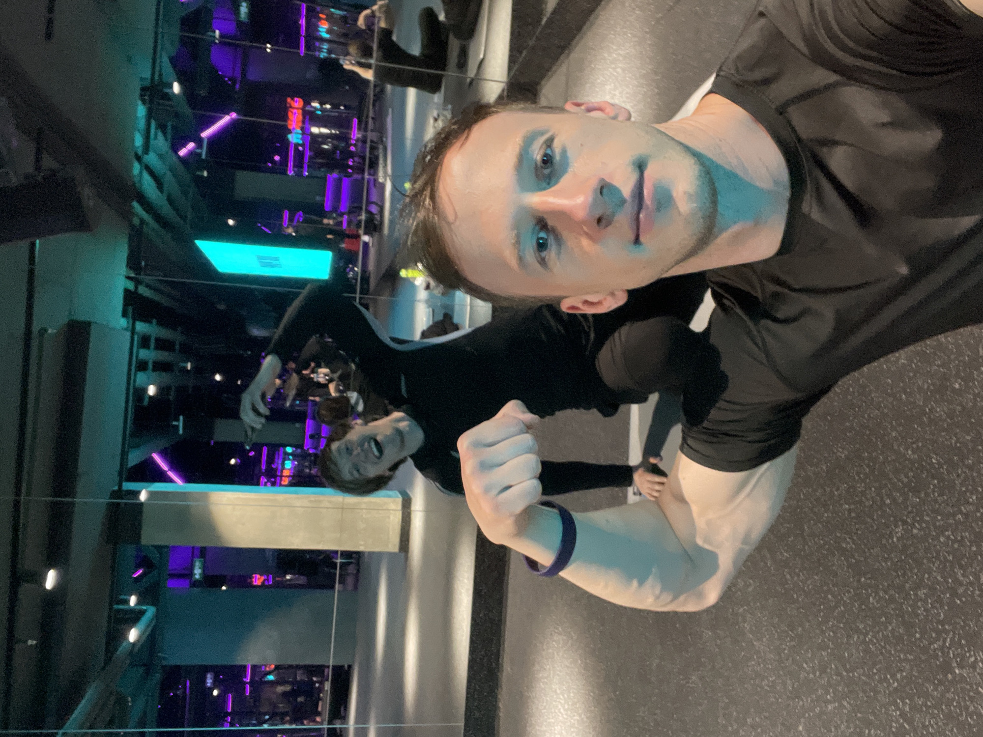 A selfie of a man in a black shirt flexing his arm. Behind him, a mirror reflects a gym setting with purple and blue neon lights. The reflection also shows another person, possibly a workout buddy, in a black outfit striking a pose. Based on the presence of the gym, the location is likely Murino, Russia.