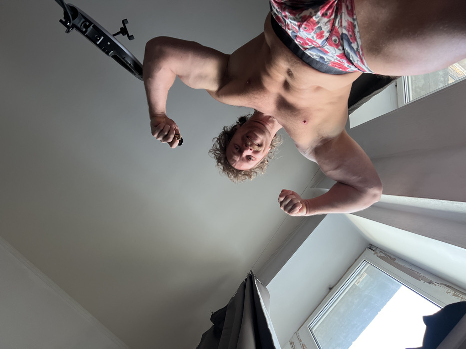 An upside-down, low-angle shot features a muscular man. He is wearing patterned underwear and flexing both arms. A ring light is visible in the upper left corner. He has short, curly hair and a mustache. A window is present on the right, while a curtain appears on the left. The ceiling dominates the background.