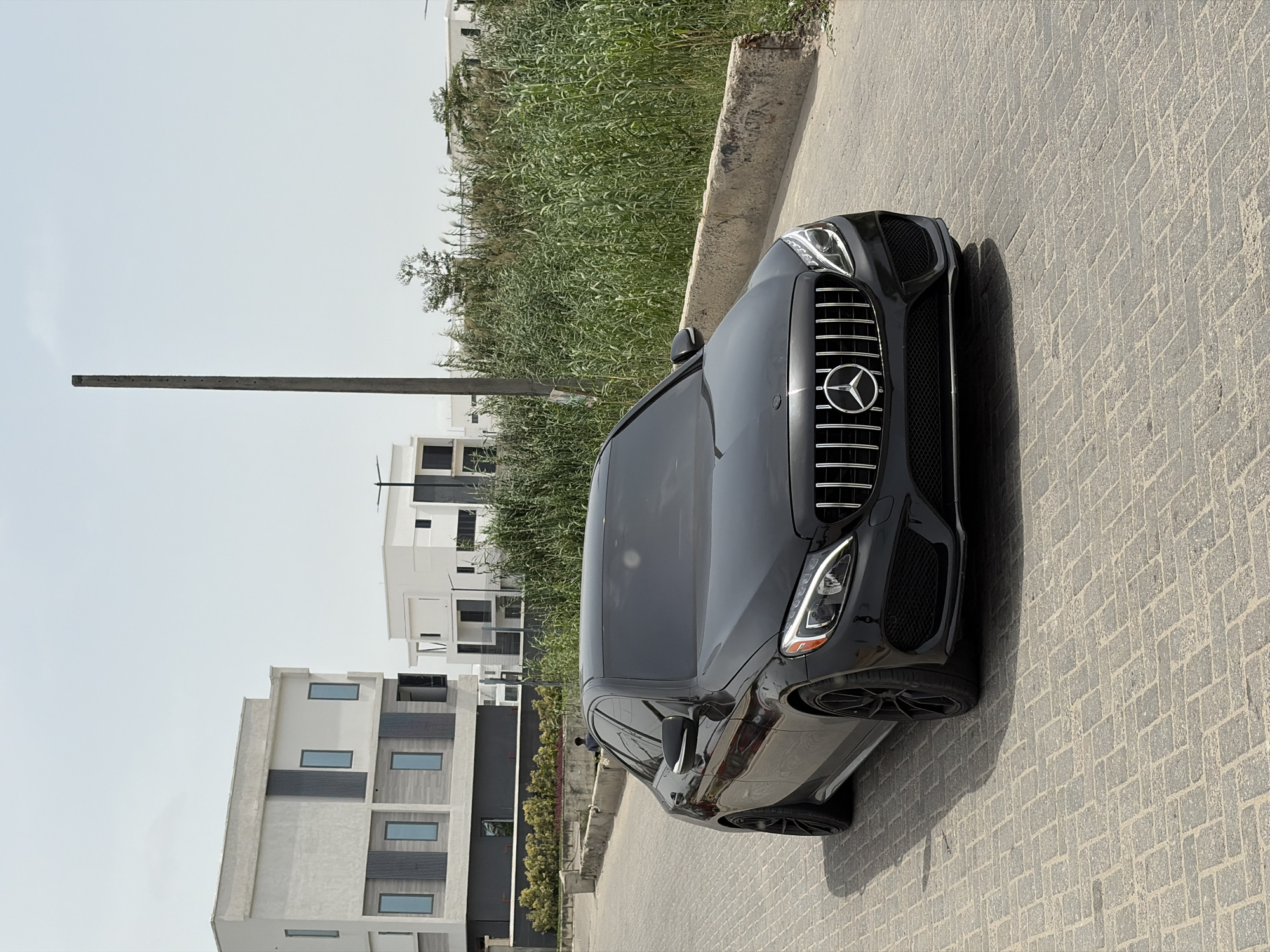 The image shows a black Mercedes-Benz vehicle parked on a paved road in Moba, Nigeria. The vehicle is positioned at an angle in the right side of the frame. In the background, there are modern residential buildings. Tall grass and a concrete wall run along the street. The sky is overcast. A concrete pole stands in the middle ground.