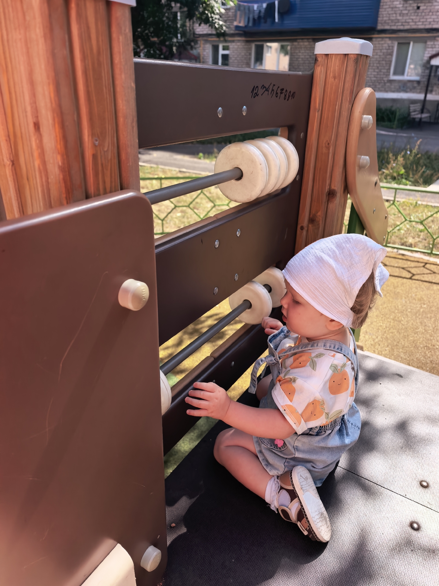 A young child, wearing a white headscarf and denim overalls over a short-sleeved shirt with an orange fruit pattern, sits on a dark-colored surface of a playground structure. The child is facing to the left, looking down at a large abacus-like toy attached to the structure. The abacus has white circular beads on metal rods. The playground structure appears to be made of wood and brown plastic. In the background, a residential building with a blue roof and windows is visible, along with some greenery and a fence. The ground surface of the playground is covered in a light brown, granular material. The scene suggests an outdoor daytime setting, likely a playground in the city of Sterlitamak, Russia.