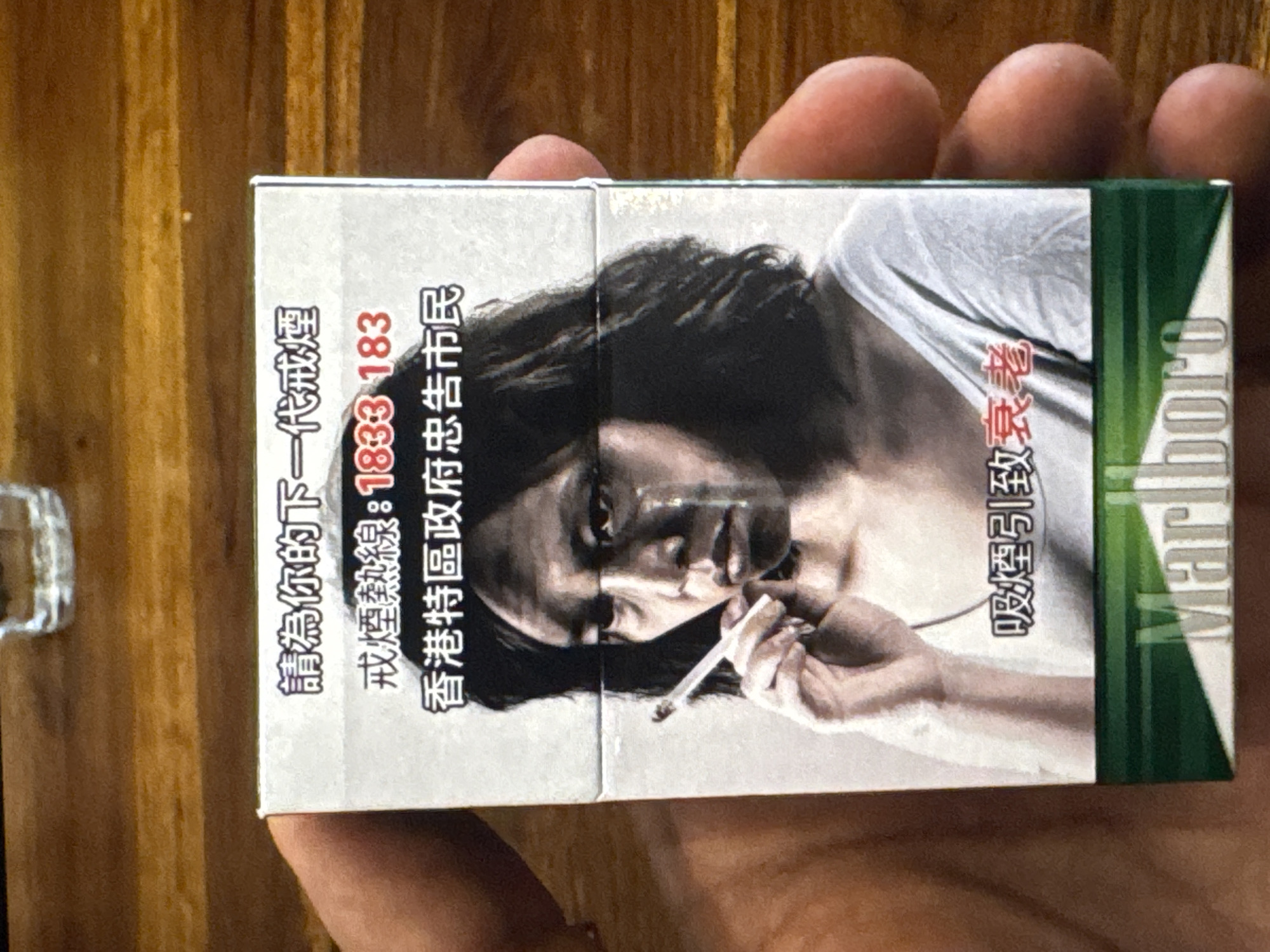 A close-up, eye-level shot shows a person's hands holding a cigarette pack with an anti-smoking message and a grim image of a woman crying while smoking. The pack is predominantly white with a green band at the bottom and features Chinese text that translates to "Please quit smoking for the next generation" and "Smoking Hotline: 1833 183". The image of the woman appears to be a painting or drawing, with dark smudges on her face and visible tears. The Marlboro logo is visible on the green band. The background is a wooden surface, possibly a table, with a blurred glass in the upper right corner. The image is sharp and well-lit, with the focus on the cigarette pack and the hands holding it. There are no identifiable landmarks or clues to suggest a specific location within Kuta Selatan, Indonesia.