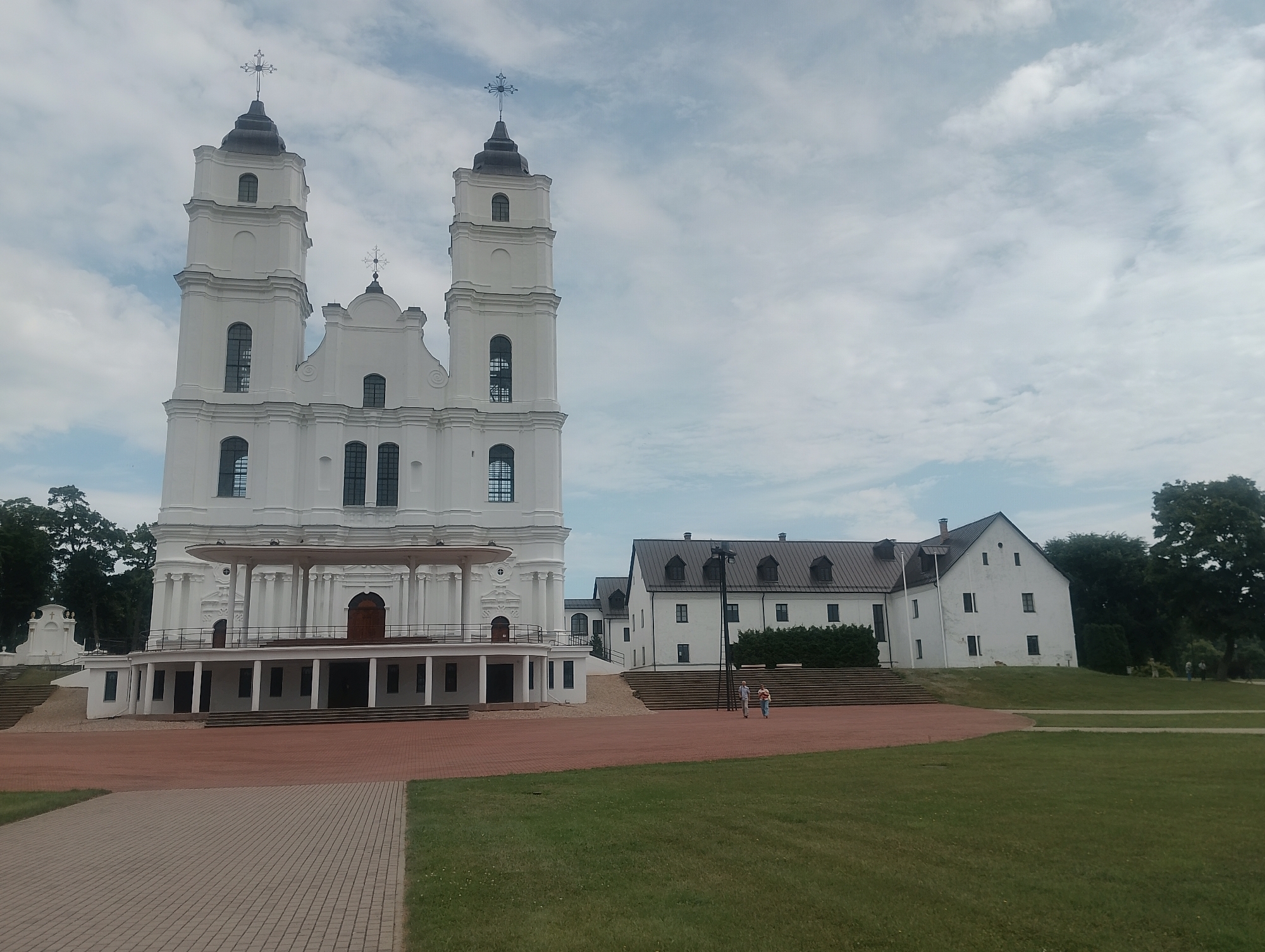 This is a wide-angle, exterior shot of the Basilica of the Assumption of the Blessed Virgin Mary in Aglona, Latvia, under a cloudy sky. The dominant subject is the large, white, neoclassical basilica with two prominent bell towers topped with crosses. To the right of the basilica, a series of connected white buildings with dark, sloped roofs and dormer windows are visible. The foreground features a paved area with red brick and a green lawn. Two individuals, a man and a woman, are walking on the paved area in front of the connected buildings. Trees line the background on the right side of the image.
