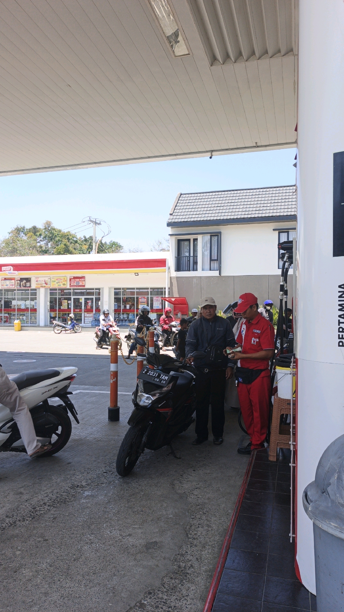 A sunny day scene unfolds at a gas station in Pangandaran, Indonesia, identified by the visible "PERTAMINA" signage on the fuel dispenser. Two men are the primary subjects of interaction. One, wearing a red uniform with "PERTAMINA" branding, appears to be a gas station attendant, handing cash to the other man, who is dressed in casual attire, including a light-colored cap. They are standing between a black motorcycle and a white scooter, which are parked on the concrete ground of the gas station forecourt.
In the background, a row of motorcycles, some with riders, suggests other customers are present. A convenience store, likely associated with the gas station, is visible with red and yellow signage. The architecture of the convenience store and a modern building with multiple windows behind it indicates an urban or semi-urban setting. The sky is clear and blue, with the sun casting shadows on the ground, typical of a bright day. The overall atmosphere suggests a routine transaction at a service station.