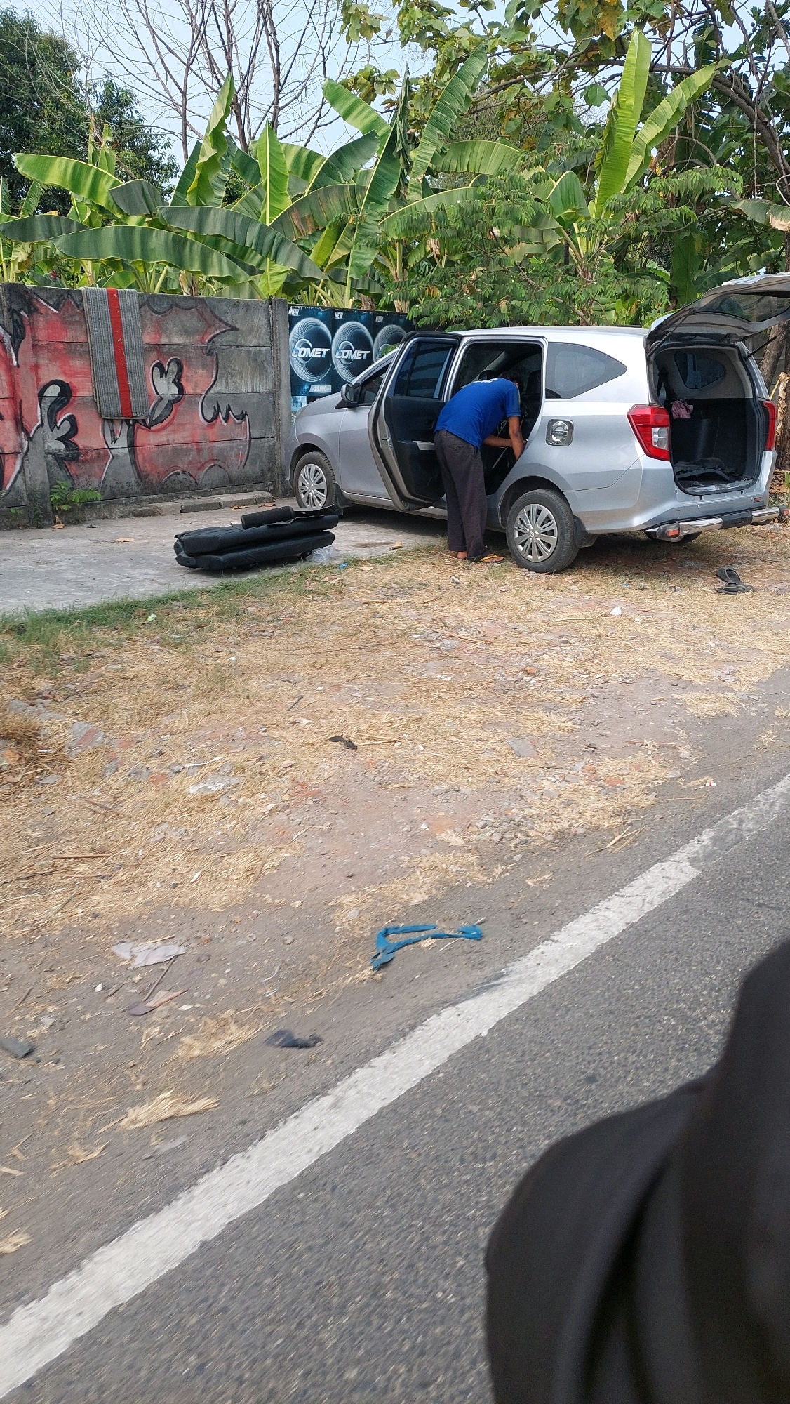 A person in a blue shirt and dark pants is bending over into the open side door of a silver minivan. The minivan has its rear hatch open, and it appears to be parked on the side of a road with a rough, dry, grassy shoulder. To the left of the minivan is a concrete wall with graffiti. In front of the minivan, on the ground, are several folded black car mats. The environment is outdoors, with trees and foliage visible in the background. The lighting suggests it is daytime.