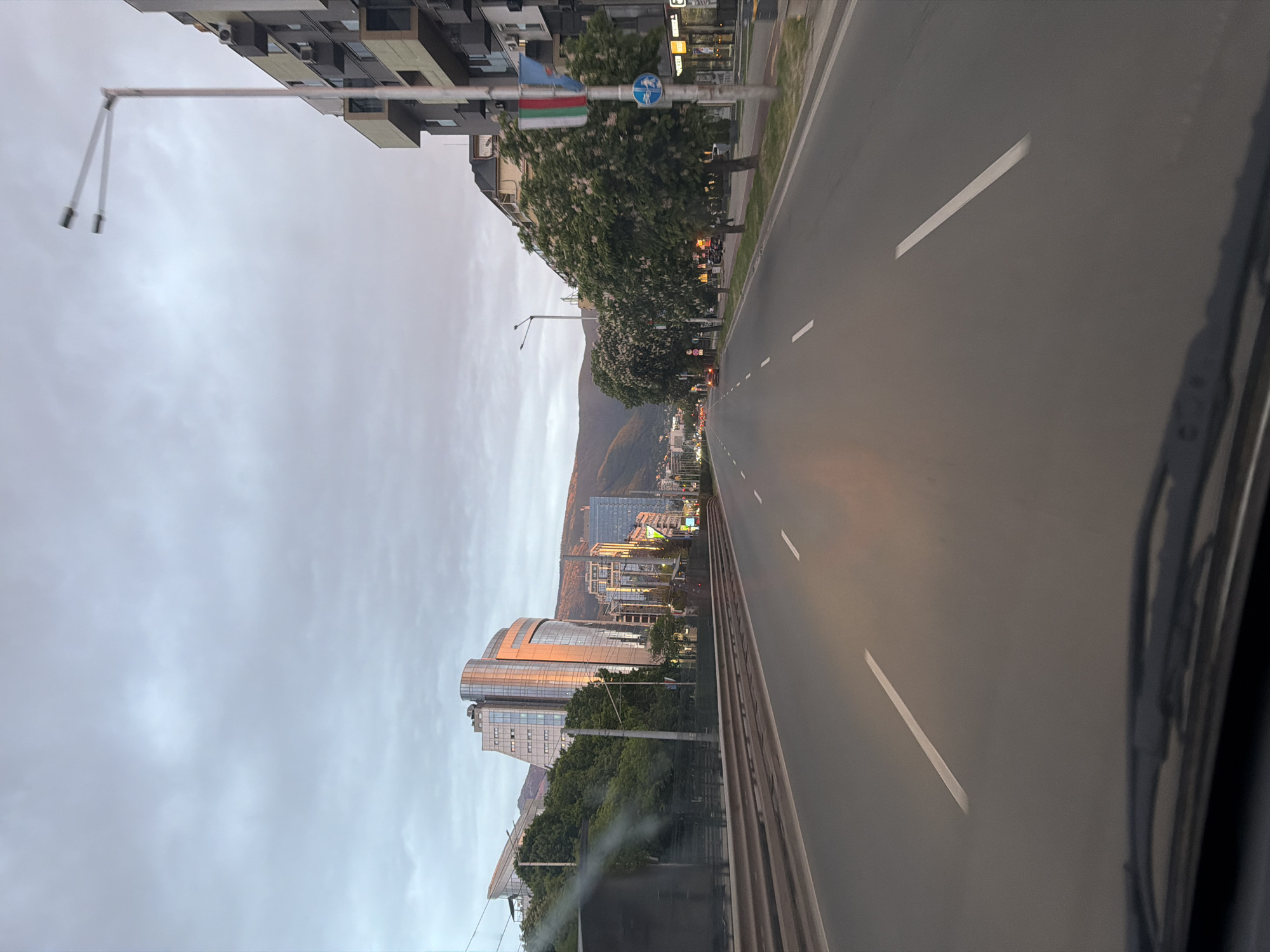 The image depicts an urban scene in Sofia, Bulgaria, likely taken from inside a moving vehicle. A straight, multi-lane road with white lane markings extends towards a cluster of tall buildings and a mountain range in the distance. A line of trees flanks the road on one side. Streetlights and signage are visible along the roadside. The sky is overcast. Buildings along the street show a mix of architectural styles. The prominent, modern buildings in the distance are located in the Mladost district of Sofia.