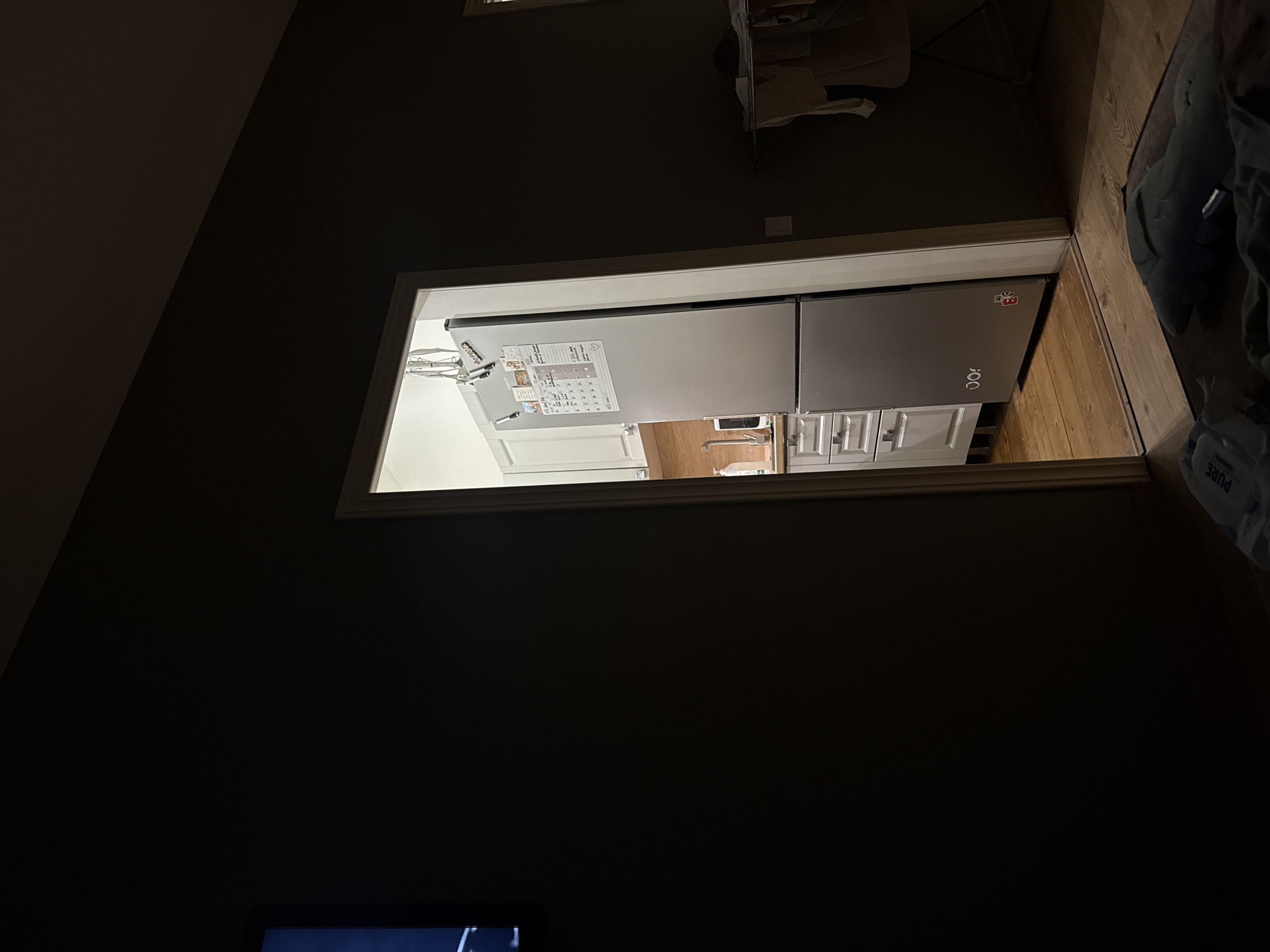 The image depicts an interior scene, likely a residential property in Рублевское Предместье Кп, Russia. The composition is dominated by a doorway leading into a kitchen area, which is partially visible. Inside the kitchen, a silver refrigerator stands against a wall, adorned with various notes and what appear to be magnets. White kitchen cabinets and drawers are also visible, along with a wooden countertop and a faucet. The floor in the kitchen is made of wooden planks.
In the foreground, outside the kitchen, the scene is darker, suggesting a living area. A portion of a television screen is visible on the left edge of the image. On the floor in the foreground, a blue fabric object resembling a blanket or cushion is present, along with some clothing items. A clothes drying rack with laundry is visible in the background, to the right of the doorway. The lighting in the image is dim, indicating either nighttime or a low-light environment. There are no people directly visible in the frame.