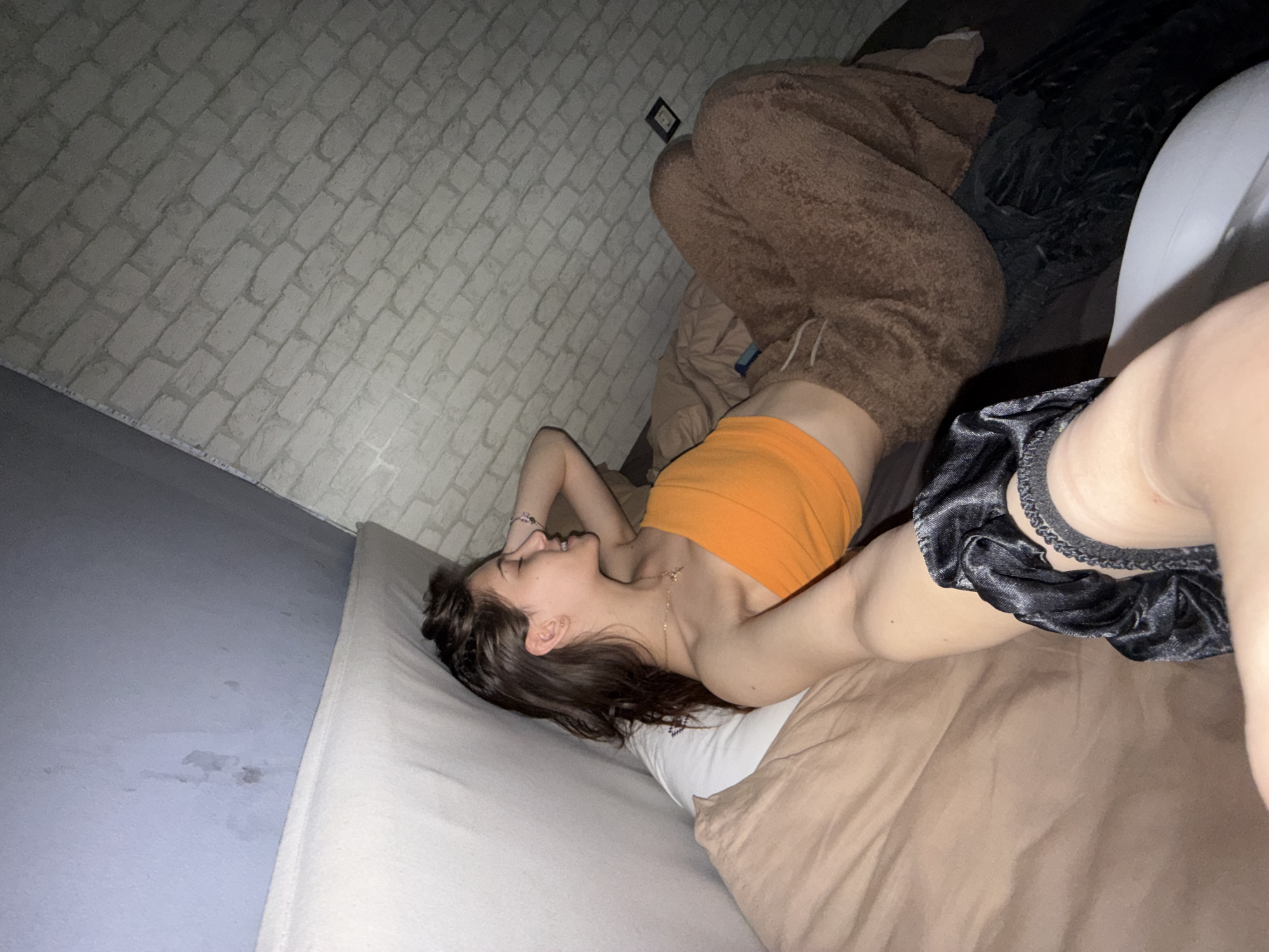 A young woman lies on a bed. She has dark brown hair, and wears an orange top and dark shorts. She appears to be in a room with white brick-patterned wallpaper. There is a teddy-bear themed pajama outfit on the bed next to her. The specific location in Sofia, Bulgaria is not discernible from the image.