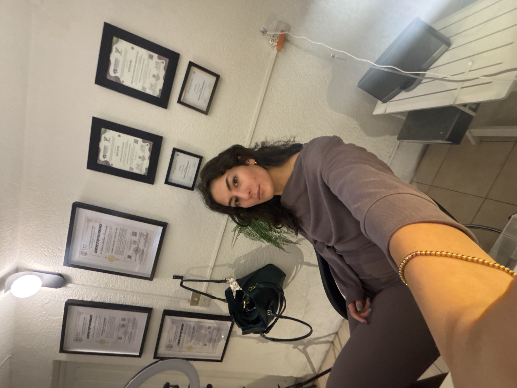 A young woman is depicted in a selfie-style indoor photograph, appearing to be seated. She has dark, wavy hair and a calm, neutral expression as she looks towards the camera. She is wearing a casual, matching two-piece outfit in a muted purplish-brown color, and a gold beaded bracelet is visible on her right wrist. Her left hand, adorned with light blue nail polish, is also in frame.
The background consists of plain white walls covered with multiple framed documents, clearly identifiable as certificates or diplomas. Several frames clearly display the words "DIPLOMA" and "CERTIFICADO," along with other Spanish text like "PROGRAMA" and "FECHA," consistent with the provided context of Puebla, Mexico. Some certificates also feature QR codes.
Other objects in the setting include a dark green handbag, embellished with a light-colored scarf, hanging on a hook mounted on the wall. A small green potted plant is visible behind the woman's head. On the left side of the image, a wall-mounted light fixture with a round bulb provides illumination, and a portion of a ring light is seen towards the bottom center, suggesting a setup for photography or video. In the upper right corner, two black speakers are placed on a white slatted shelf, with wires running across the wall. The floor is tiled with light-colored squares. The scene portrays a functional and potentially professional indoor space.