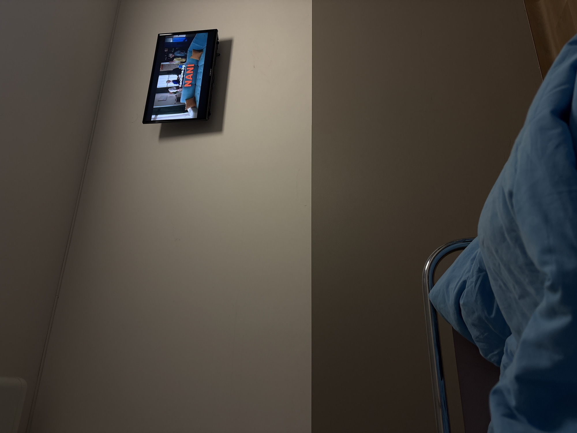 The image depicts an interior space, likely a hospital room or similar environment, with a wall-mounted television displaying a program featuring the text "NANIhome.org". To the right, part of a metal bed frame and a blue blanket or sheet are visible. The scene is neutrally lit, giving the walls a grayish-beige color. The precise location within the city of Sofia, Bulgaria, is not discernible from the visual data provided.