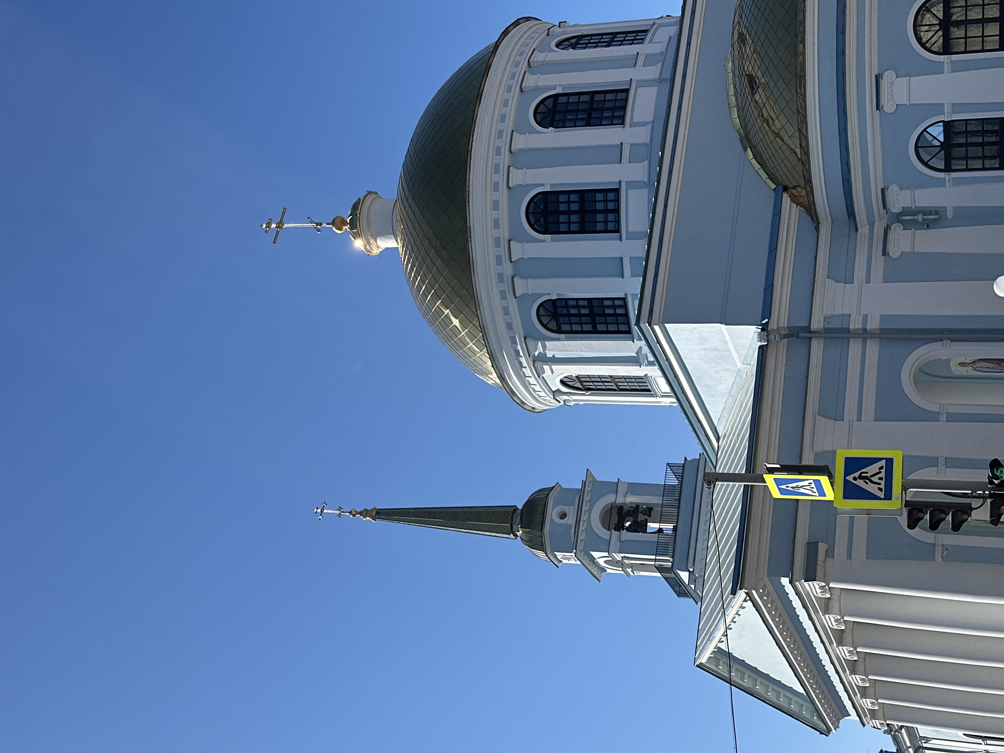 The image shows a light blue and white building with golden domes against a clear blue sky. The building's architecture is reminiscent of a Russian Orthodox church. A cross sits atop the taller of the two domes visible. In the foreground, a pedestrian crossing sign is mounted on a pole with a traffic light. The location is likely the city of Votkinsk, Russia.