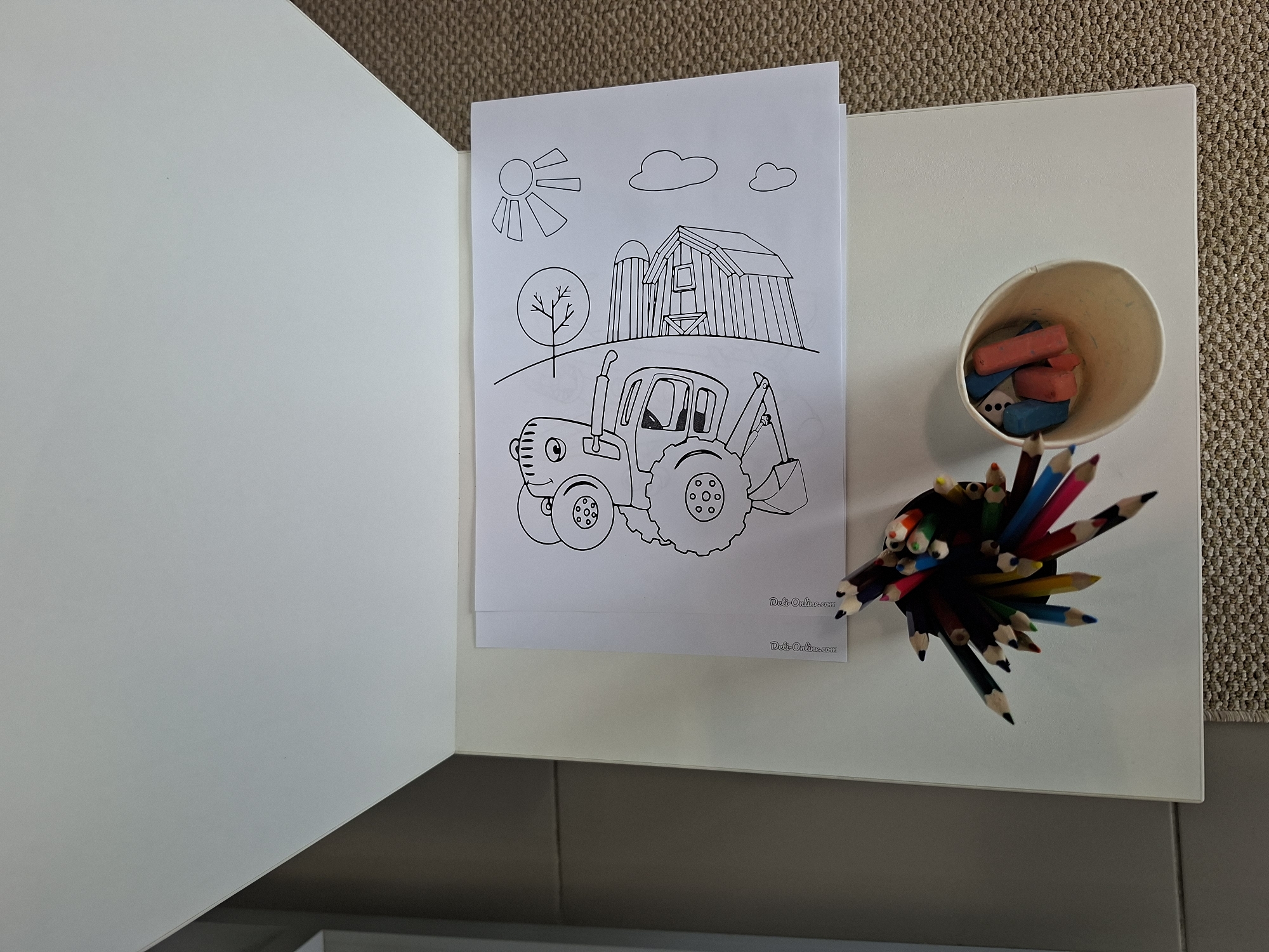 A coloring book with a picture of a tractor, a barn, a tree, the sun, and clouds is on a white surface. A collection of colored pencils is in a cup next to the coloring book. There are also some chalk sticks in the cup. The scene appears to be indoors, possibly on a table or desk, with a carpeted floor visible in the background. No people are present in the image. The location in Izhevsk, Russia cannot be determined from the image.
