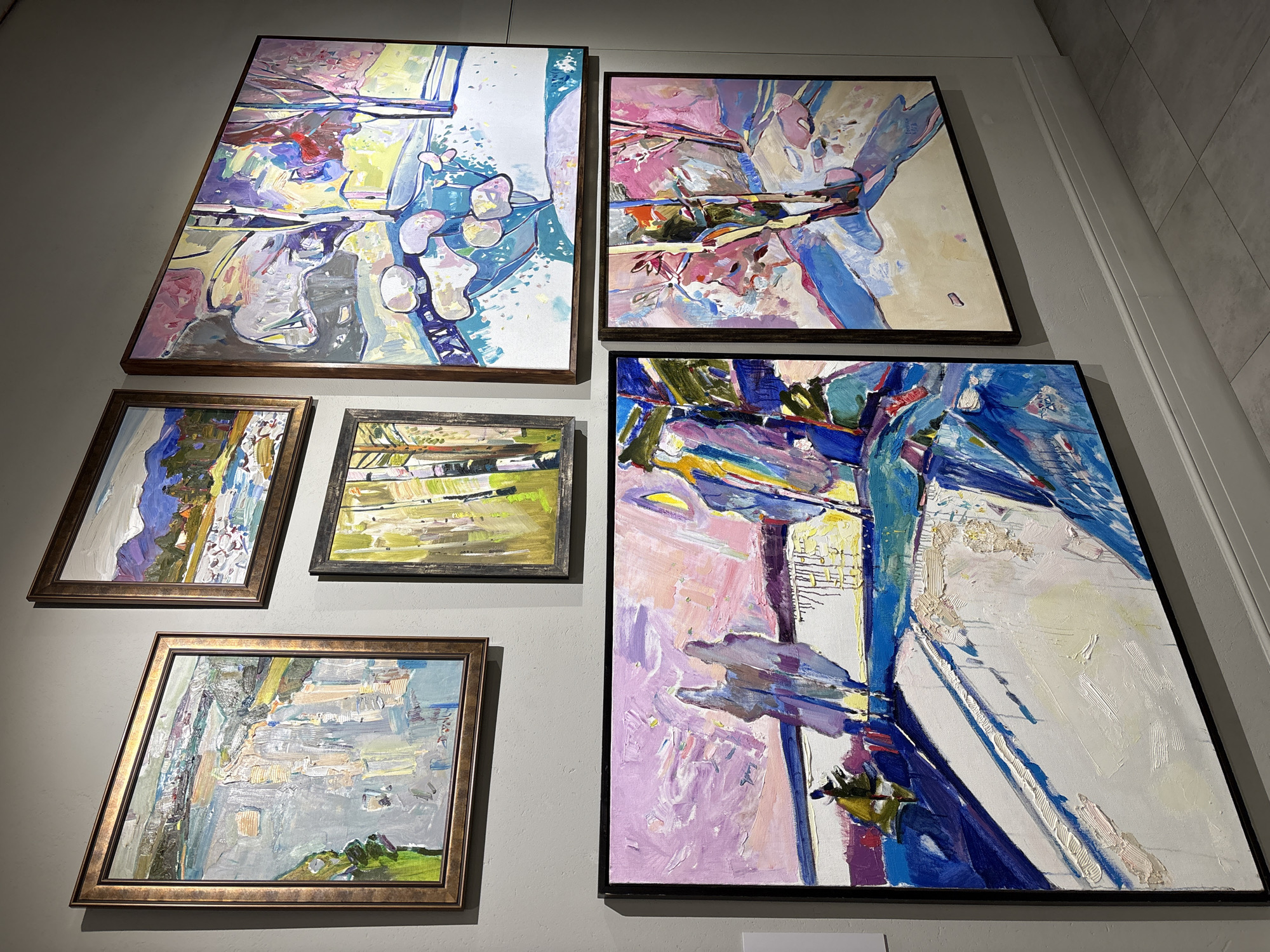 A wall in what appears to be a gallery or exhibit space displays six framed paintings. The two largest paintings are situated on the upper right and lower right, while the remaining four, smaller paintings are on the left. The paintings feature abstract, colorful depictions with broad brushstrokes. The gallery is located in Irkutsk, Russia.