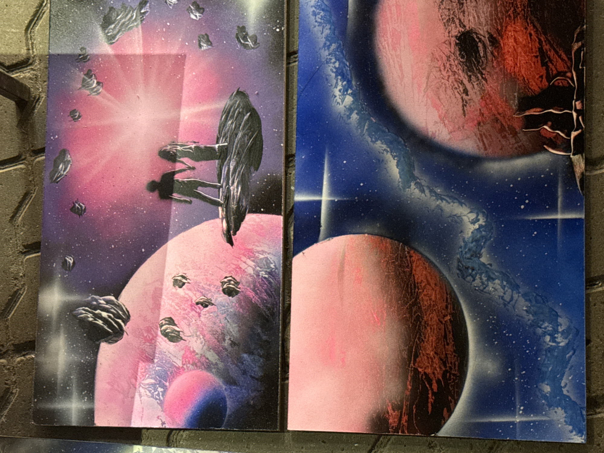 This is a media file featuring two spray-painted artworks depicting a space scene. The left panel shows a vibrant nebula with pink and purple hues, a bright white light source emanating rays, and several dark, irregular shapes that appear to be asteroids or debris. Two silhouetted figures are visible in the foreground, seemingly reaching out or interacting with a large, dark, textured object that resembles a spaceship or a strange celestial body. There are also smaller, dark, pointed objects scattered throughout the scene, possibly also spaceships or asteroids. A large, textured pink planet is prominent in the lower part of this panel, with a smaller, glowing purple and blue sphere beneath it.
The right panel presents a different view of space, dominated by deep blues and speckled white stars. Two large planets are depicted; one is a predominantly pink sphere with dark, textured markings suggesting a rough surface. The other planet, partially visible on the right edge, appears red and intricately textured. A swirling nebula or cosmic cloud in shades of blue and white is visible between the planets. Two bright, star-like flares are present in the lower right section.
The artworks convey a sense of wonder and exploration, characteristic of science fiction themes. The spray-paint technique creates a dreamy, otherworldly atmosphere. There is no visible text within the images. The context suggests these are likely displayed in a public space in Kazan, Russia, possibly as art pieces. The time of day and weather are irrelevant as it is an artistic representation of space.