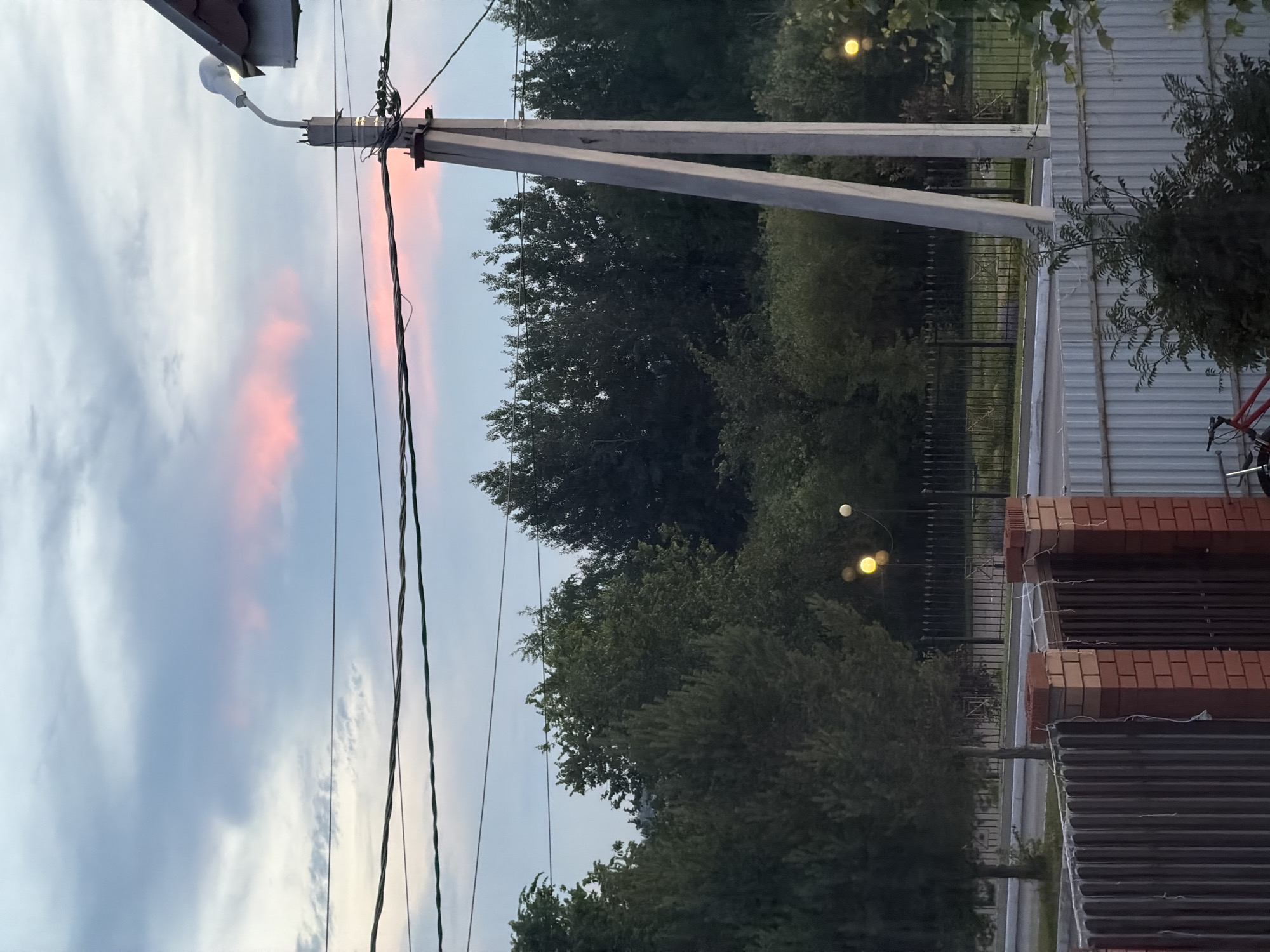 The image depicts an outdoor scene with a twilight sky featuring pink and blue clouds. In the foreground, there is a brick and metal fence. Behind the fence, power lines stretch across the frame, supported by a concrete utility pole with a street lamp. A line of trees forms a dark silhouette against the sky, with some scattered greenery and a darker forested area further back. Two yellow streetlights are visible in the middle ground, illuminating a fenced-off area. A portion of a red bicycle is visible at the bottom left corner of the image. The overall impression is a suburban or rural setting at dusk.