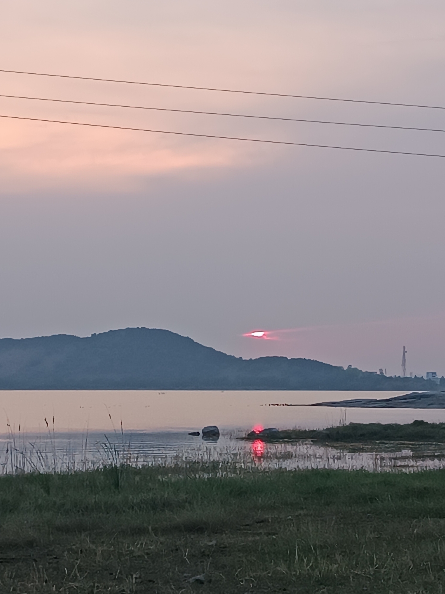 The image depicts a landscape scene in Kammavaripalli, India, at twilight. In the foreground, there's grassy vegetation leading to a body of water, possibly a lake or reservoir. The water reflects a faint, reddish glow from the partially obscured setting sun, which is positioned just above the horizon. A low, rolling hill or mountain is visible in the mid-ground, silhouetted against the fading sky. The sky is overcast with a muted pinkish-gray hue. Electrical wires are strung across the upper portion of the frame. In the distance, on the right side of the image, are buildings, and what appears to be a tall antenna or tower. The overall scene is tranquil and natural, with minimal signs of human activity.