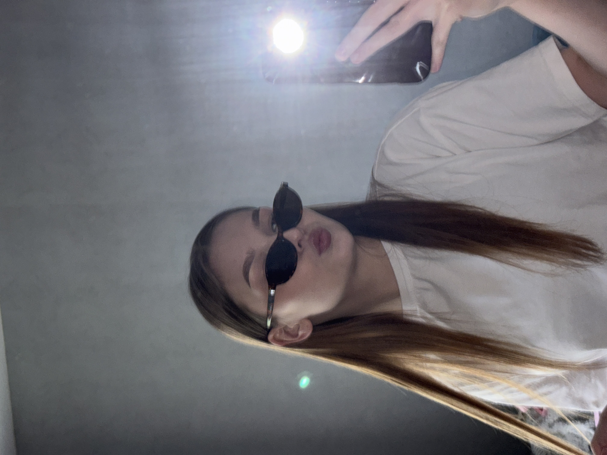 A young woman wearing sunglasses and a white shirt is taking a selfie. She is holding a phone with a bright flash on. The background is a plain, light-colored wall. The location is not discernible as Chaikovsky, Russia.